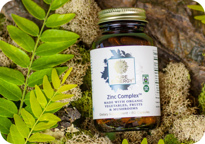 A bottle of Pure Synergy Zinc Complex organic capsules rests on green leaves and moss.