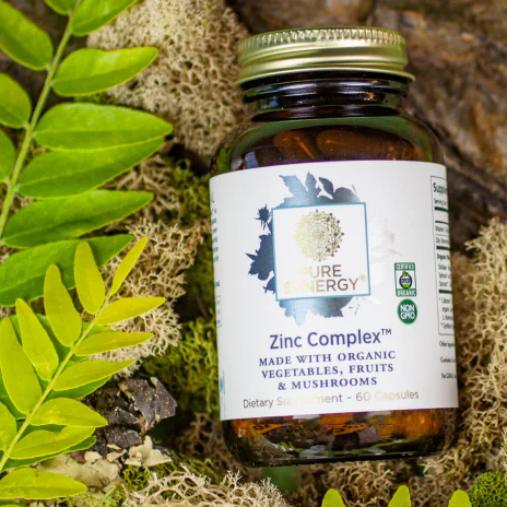 A bottle of Pure Synergy Zinc Complex organic capsules rests on green leaves and moss.