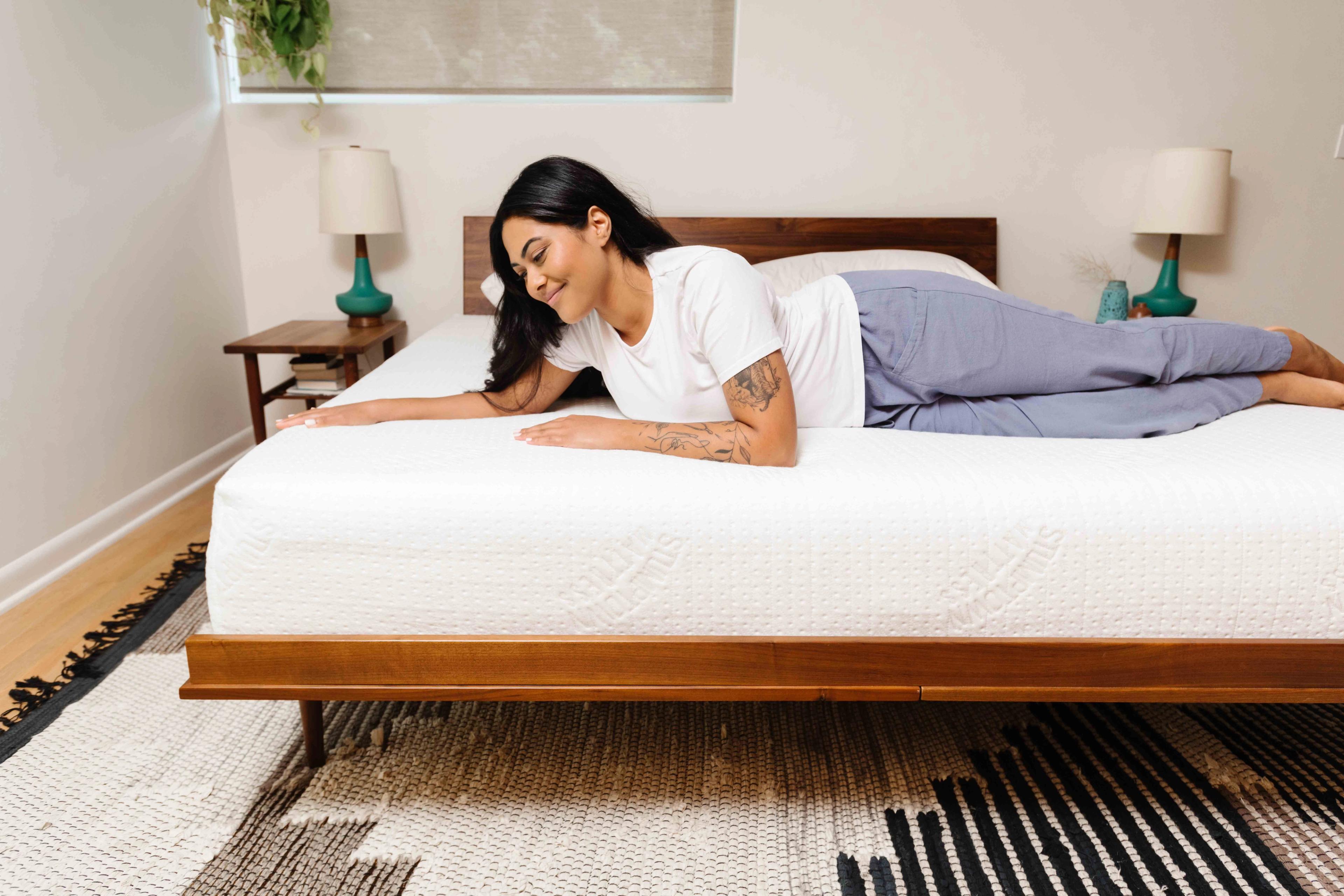 A smiling woman lies comfortably on a white mattress, her hand resting on its surface.