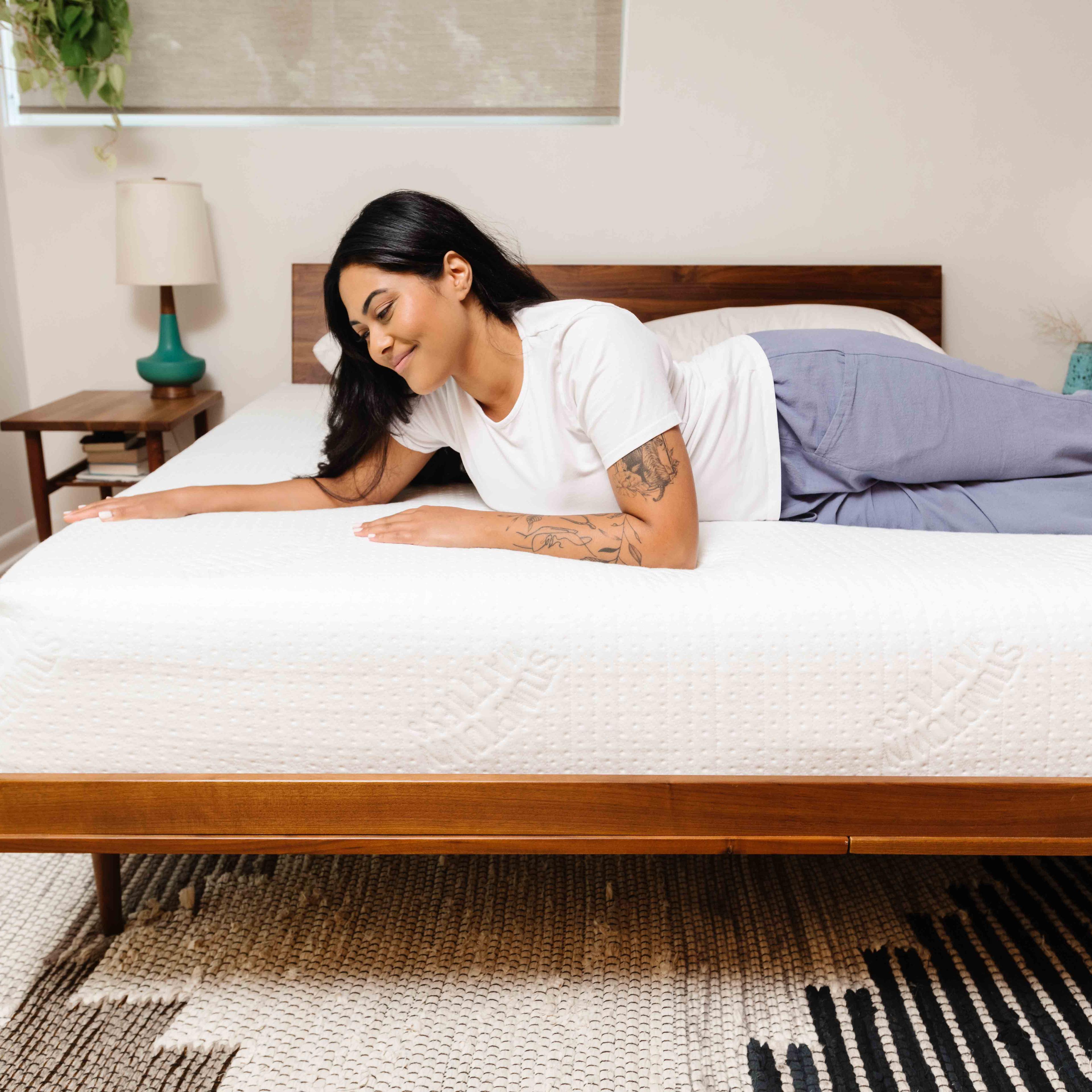 A smiling woman lies comfortably on a white mattress, her hand resting on its surface.