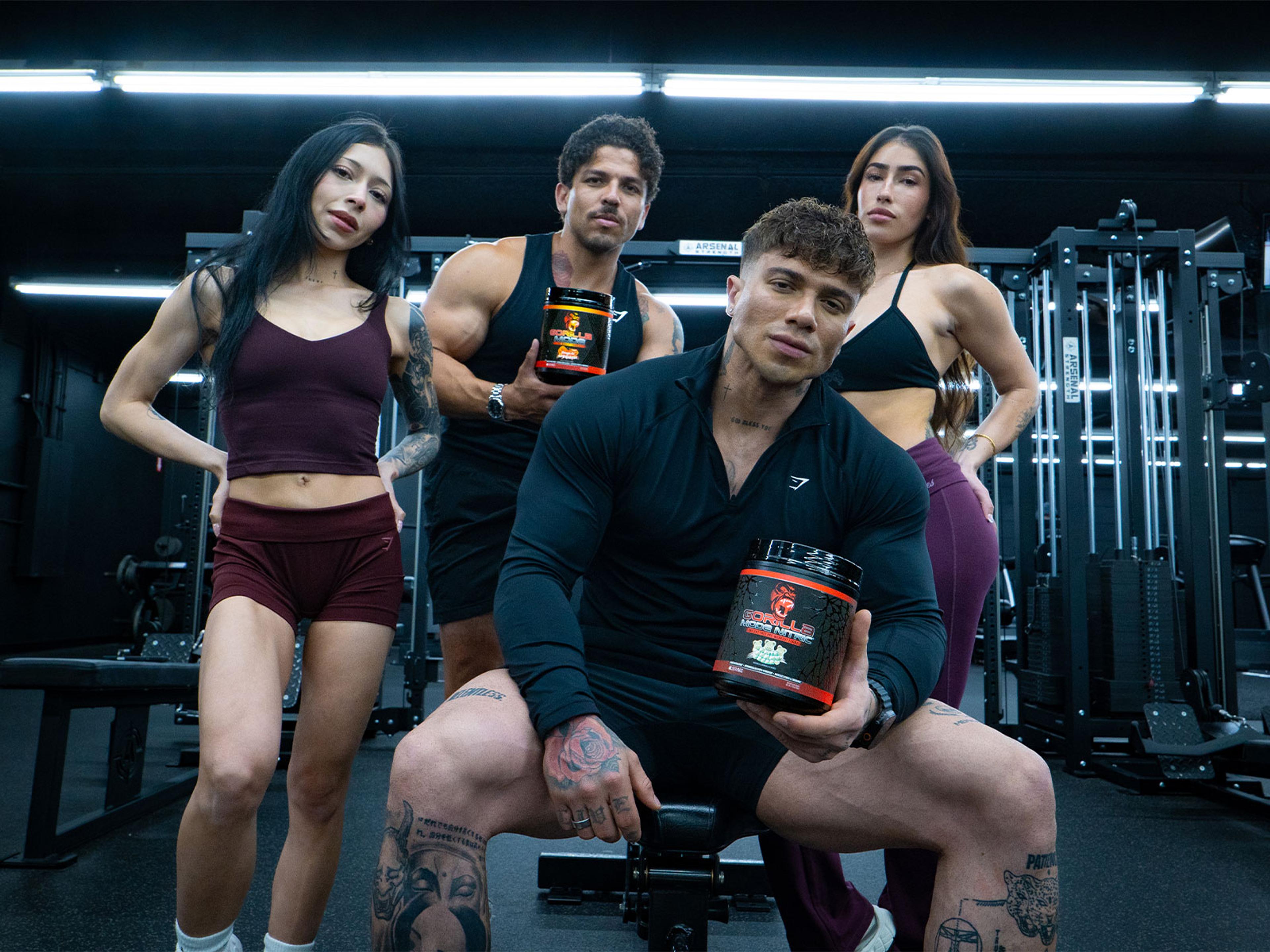 Four fitness models, two men and two women, pose in a gym, with two holding black "Gorilla Supps" supplement tubs.