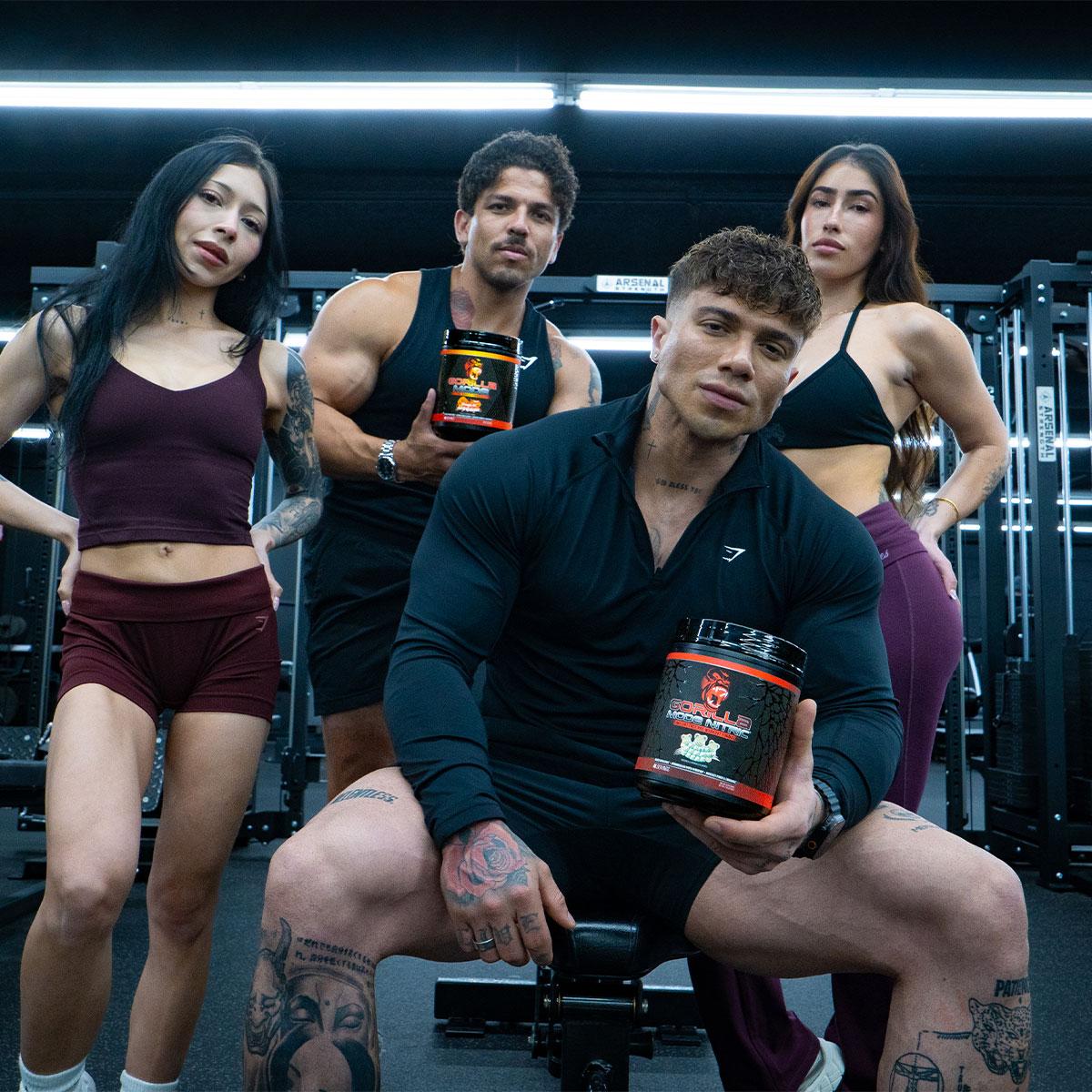 Four fitness models, two men and two women, pose in a gym, with two holding black "Gorilla Supps" supplement tubs.