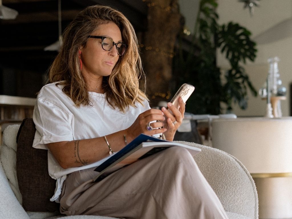 A woman wearing glasses sits in a chair, looking at her phone with a book on her lap.