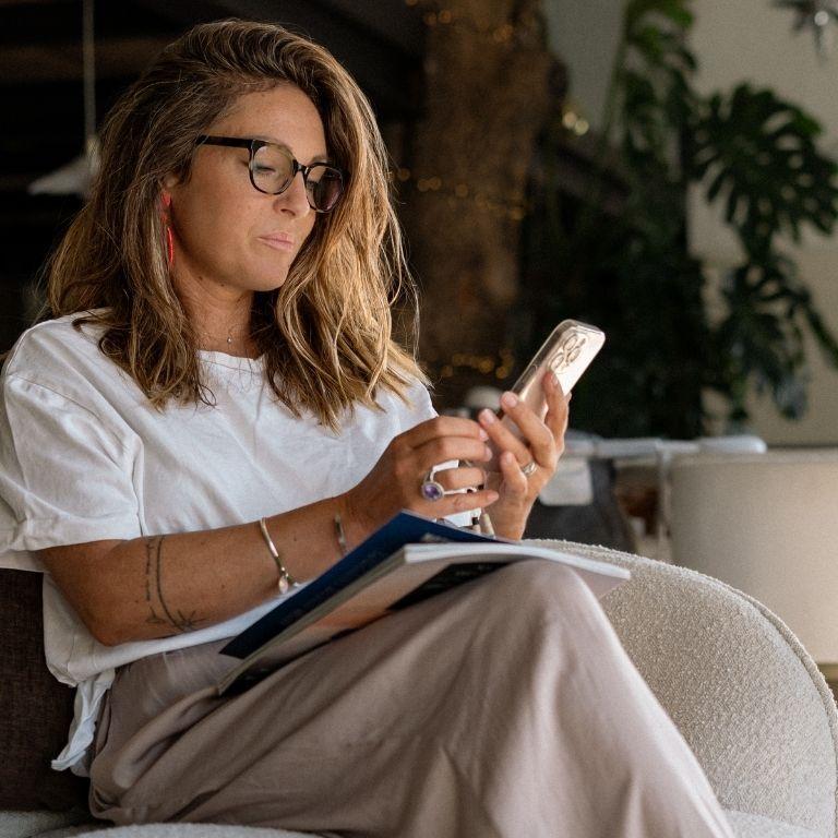 A woman wearing glasses sits in a chair, looking at her phone with a book on her lap.