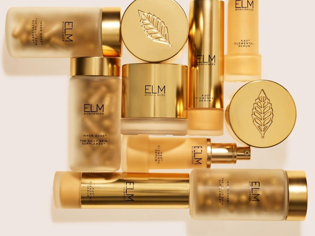 ELM BIOSCIENCES daily skin supplement, elemental serum, and night cream, in gold and frosted containers, with a separate gold leaf-embossed cap.