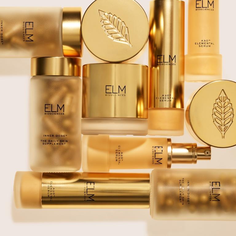 ELM BIOSCIENCES daily skin supplement, elemental serum, and night cream, in gold and frosted containers, with a separate gold leaf-embossed cap.