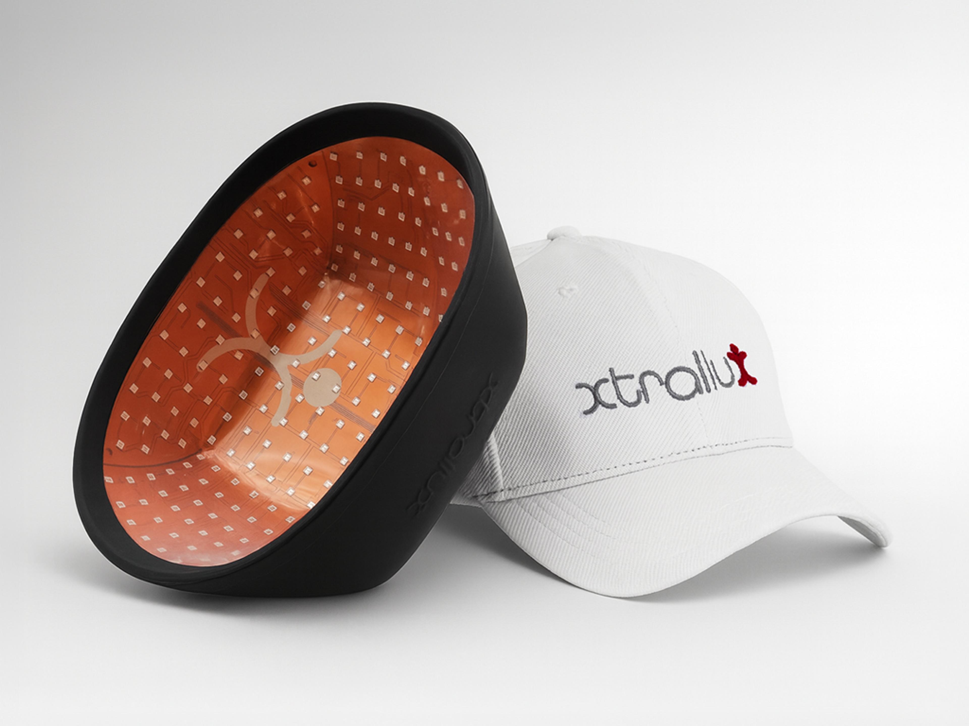 A black LED light therapy device with an orange interior next to a white Xtralux branded baseball cap.