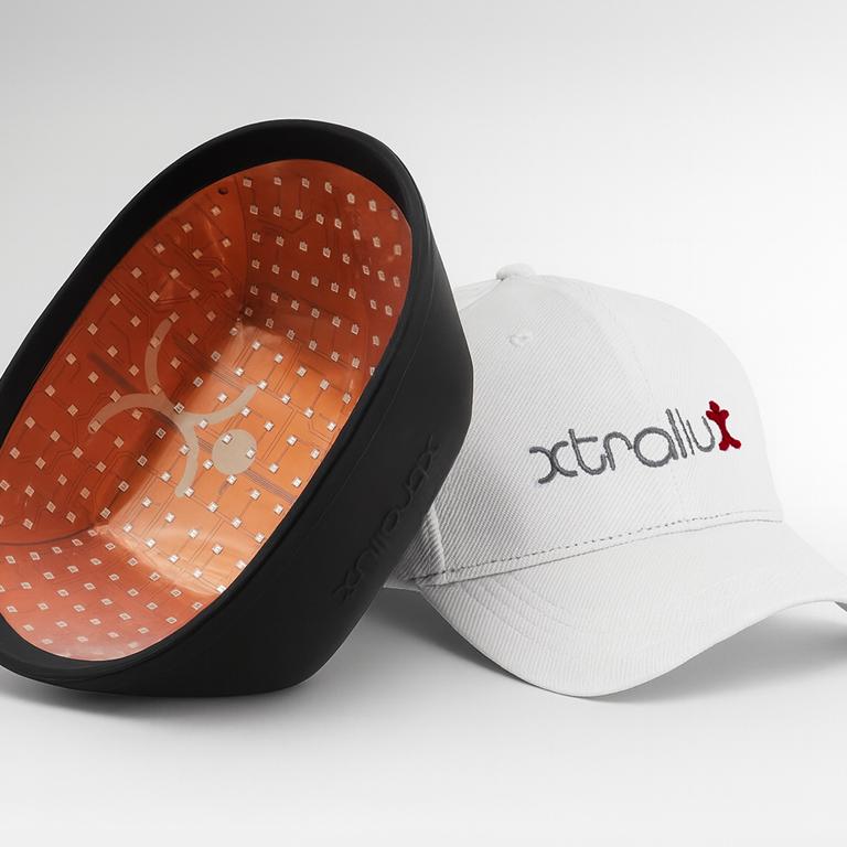 A black LED light therapy device with an orange interior next to a white Xtralux branded baseball cap.