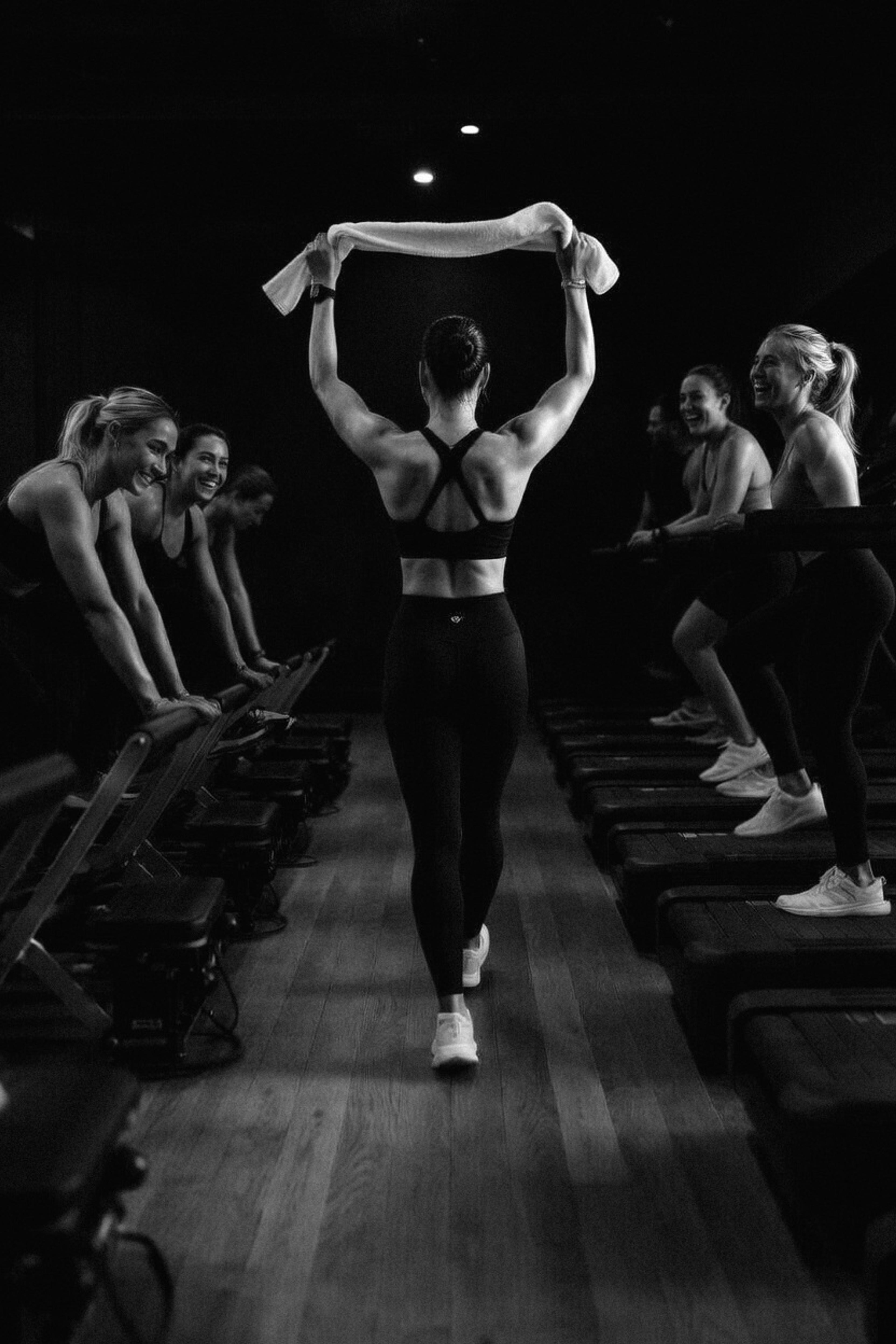 Black and white photo of a muscular woman holding a towel overhead, walking away in a gym with smiling women on fitness equipment.