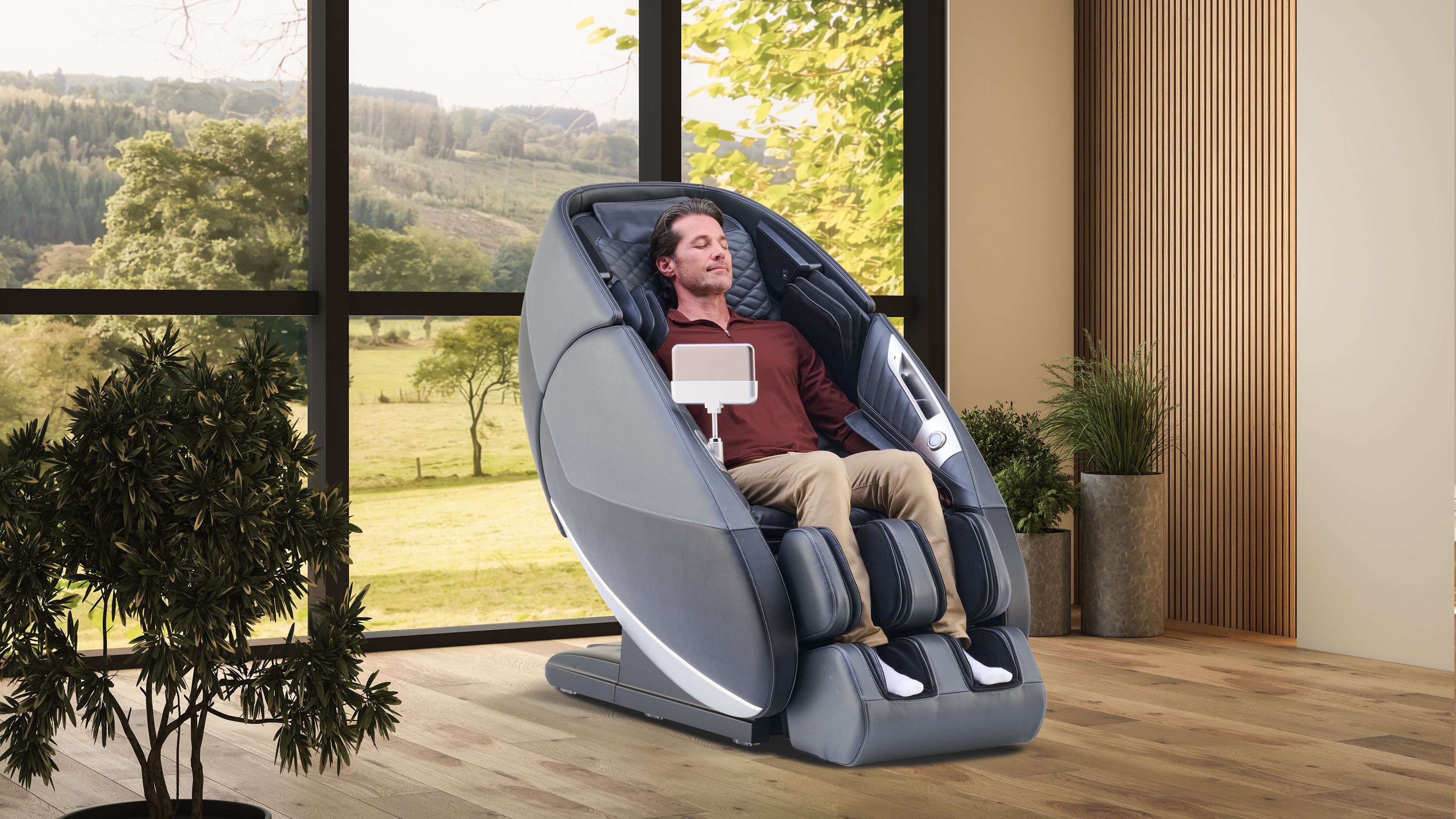 A man with closed eyes relaxes in a modern grey massage chair with a tablet, by a large window overlooking a green landscape.