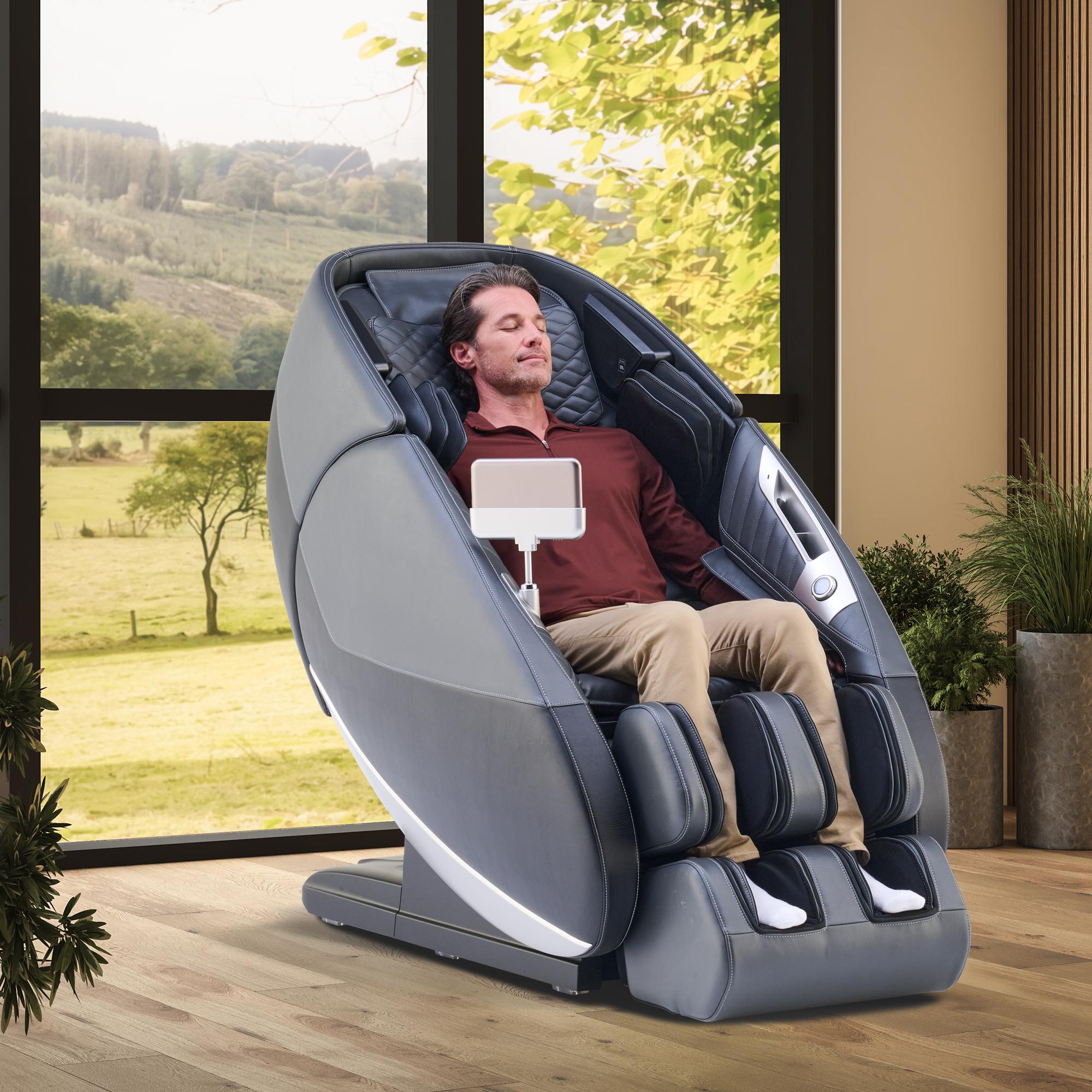 A man with closed eyes relaxes in a modern grey massage chair with a tablet, by a large window overlooking a green landscape.