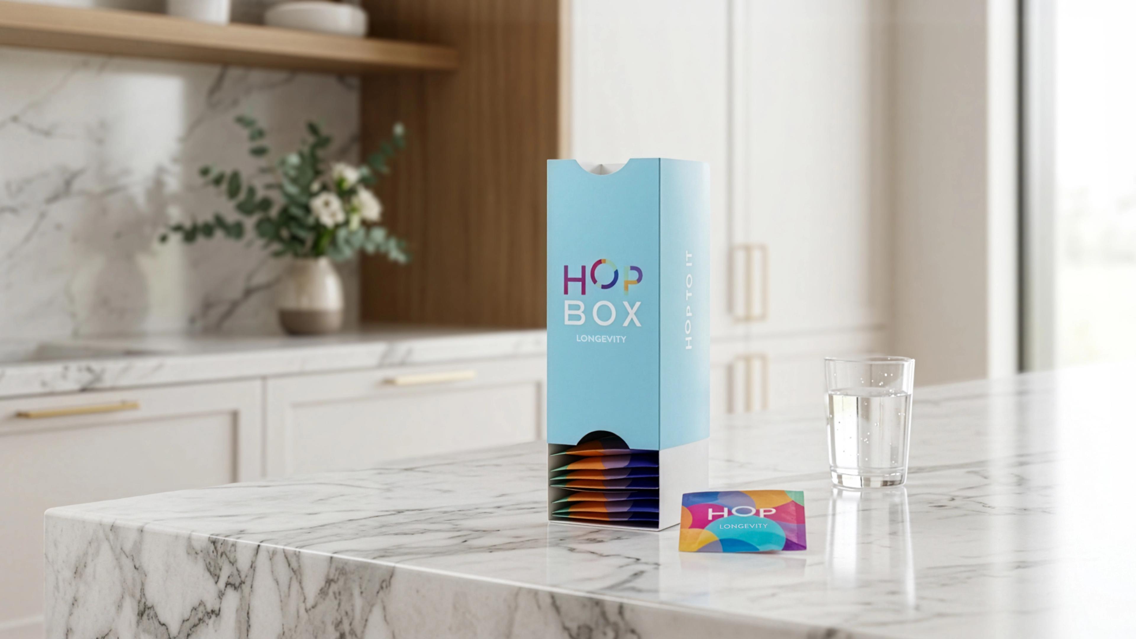 A light blue "HOP BOX LONGEVITY" dispenser with colorful packets, a single packet, and a glass of water on a marble kitchen counter.