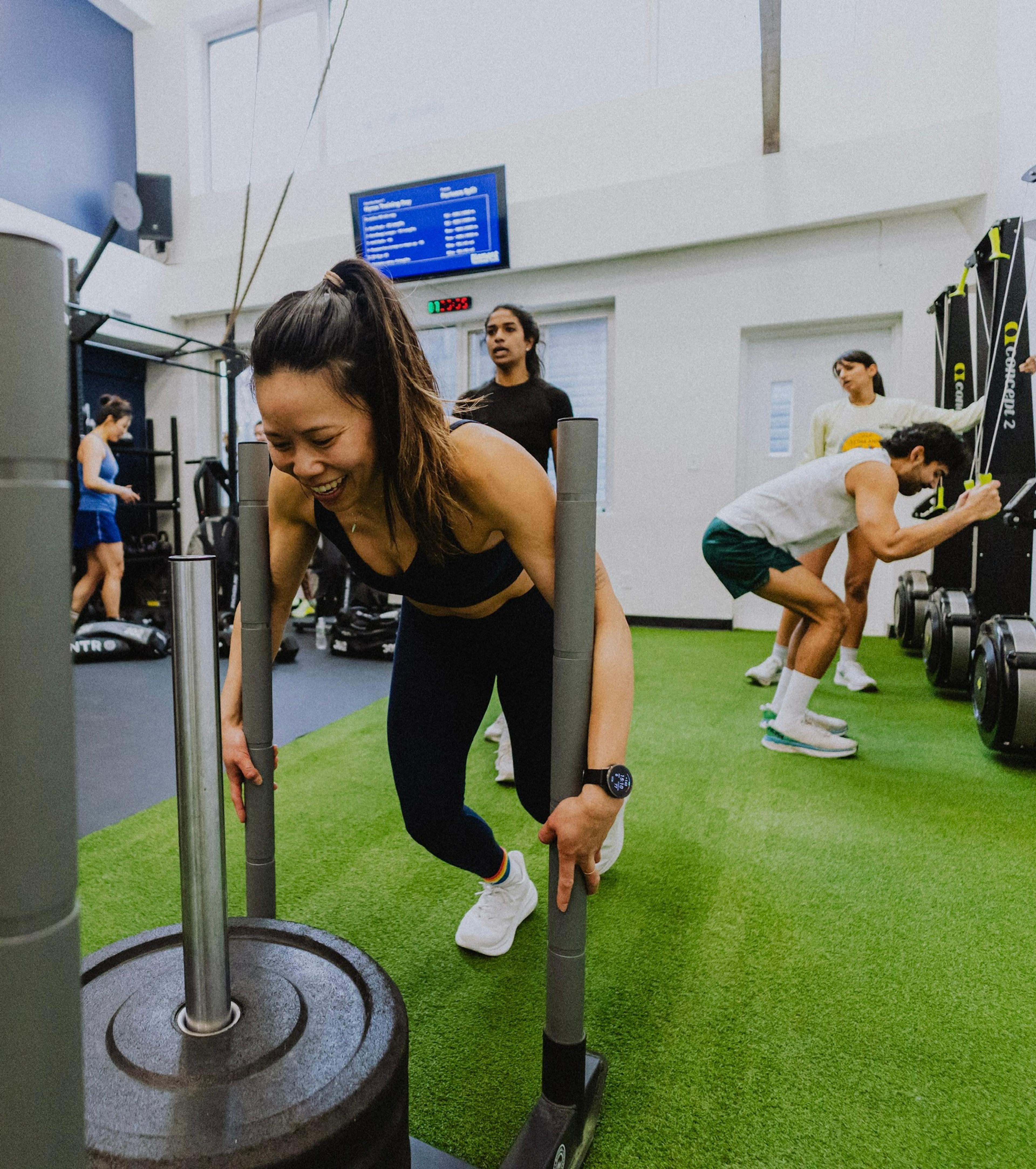 A smiling woman pushes a weighted sled on green turf in a gym.