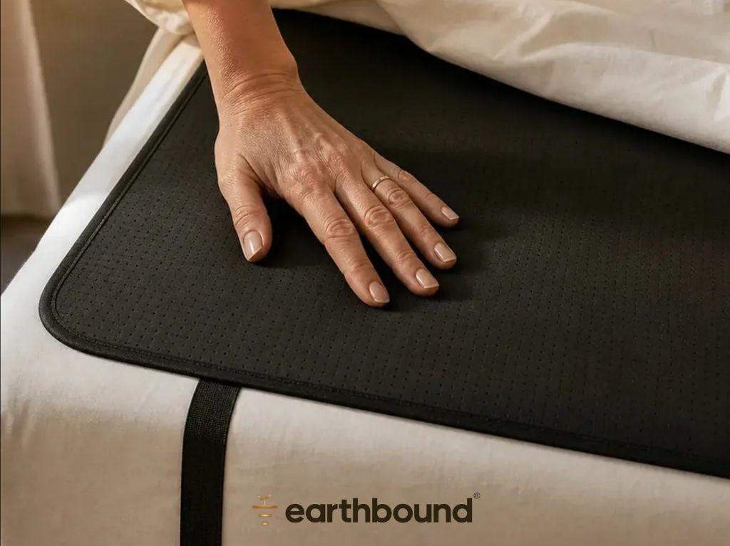 A hand rests on a black perforated mat with an 'earthbound' logo, placed on white bed sheets.