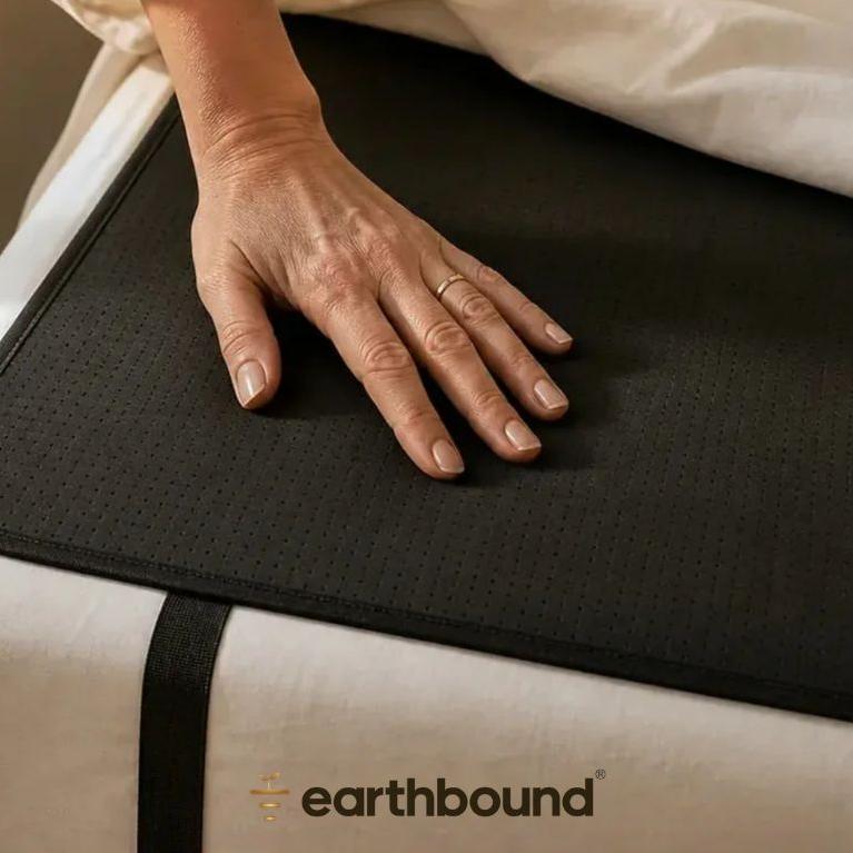 A hand rests on a black perforated mat with an 'earthbound' logo, placed on white bed sheets.