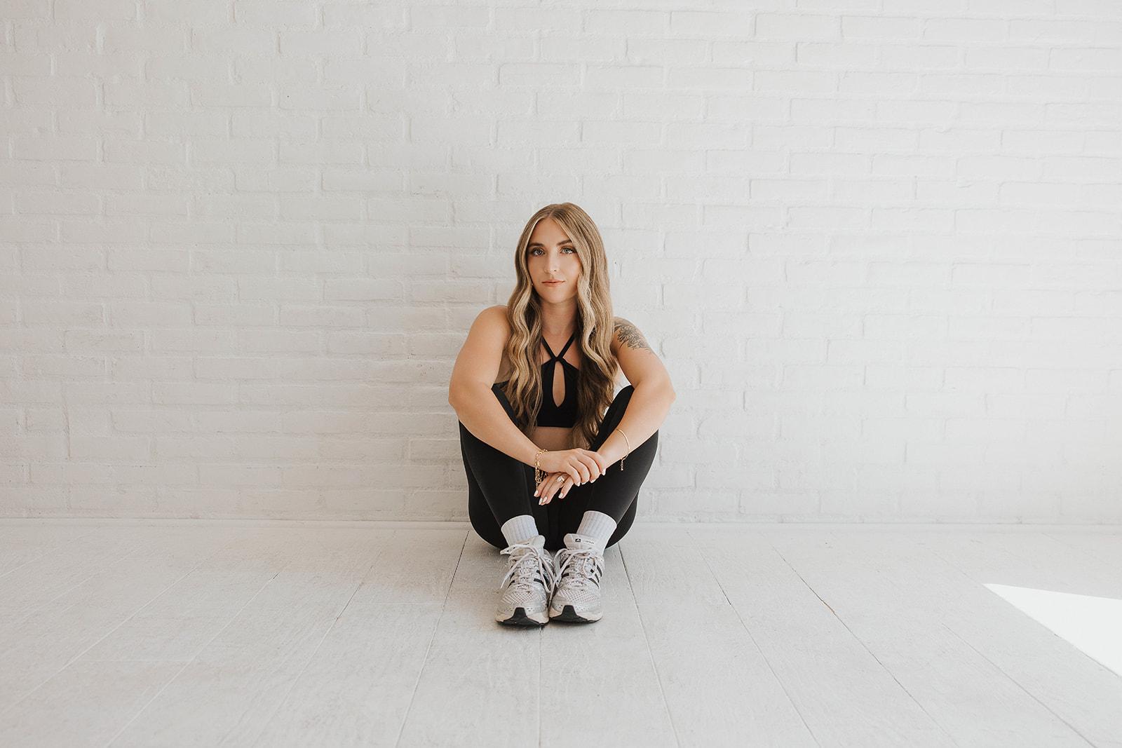 A woman in athletic wear sits on the floor against a white brick wall, looking at the camera.