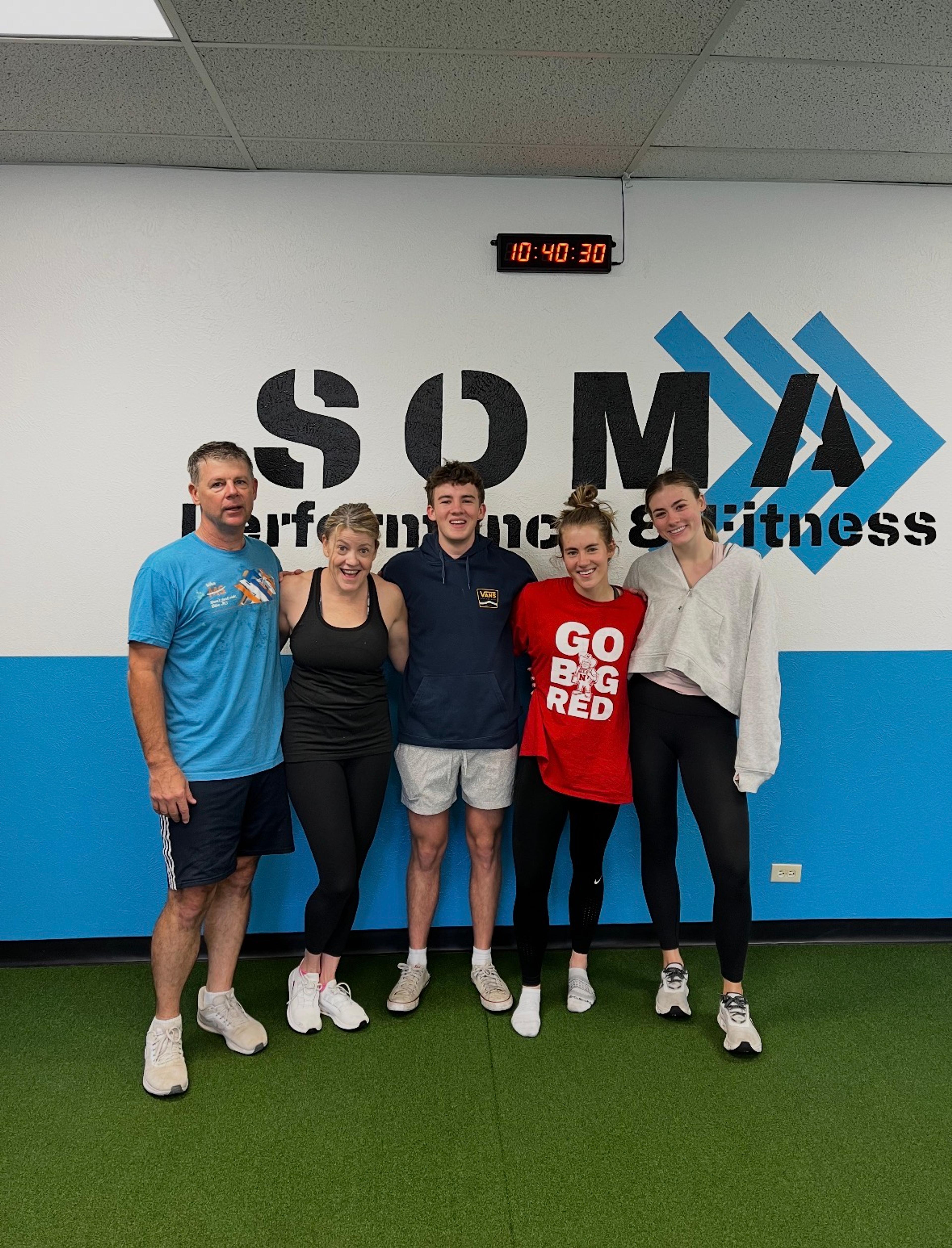 Five smiling people stand together in a gym with "SOMA Performance & Fitness" on the wall behind them.