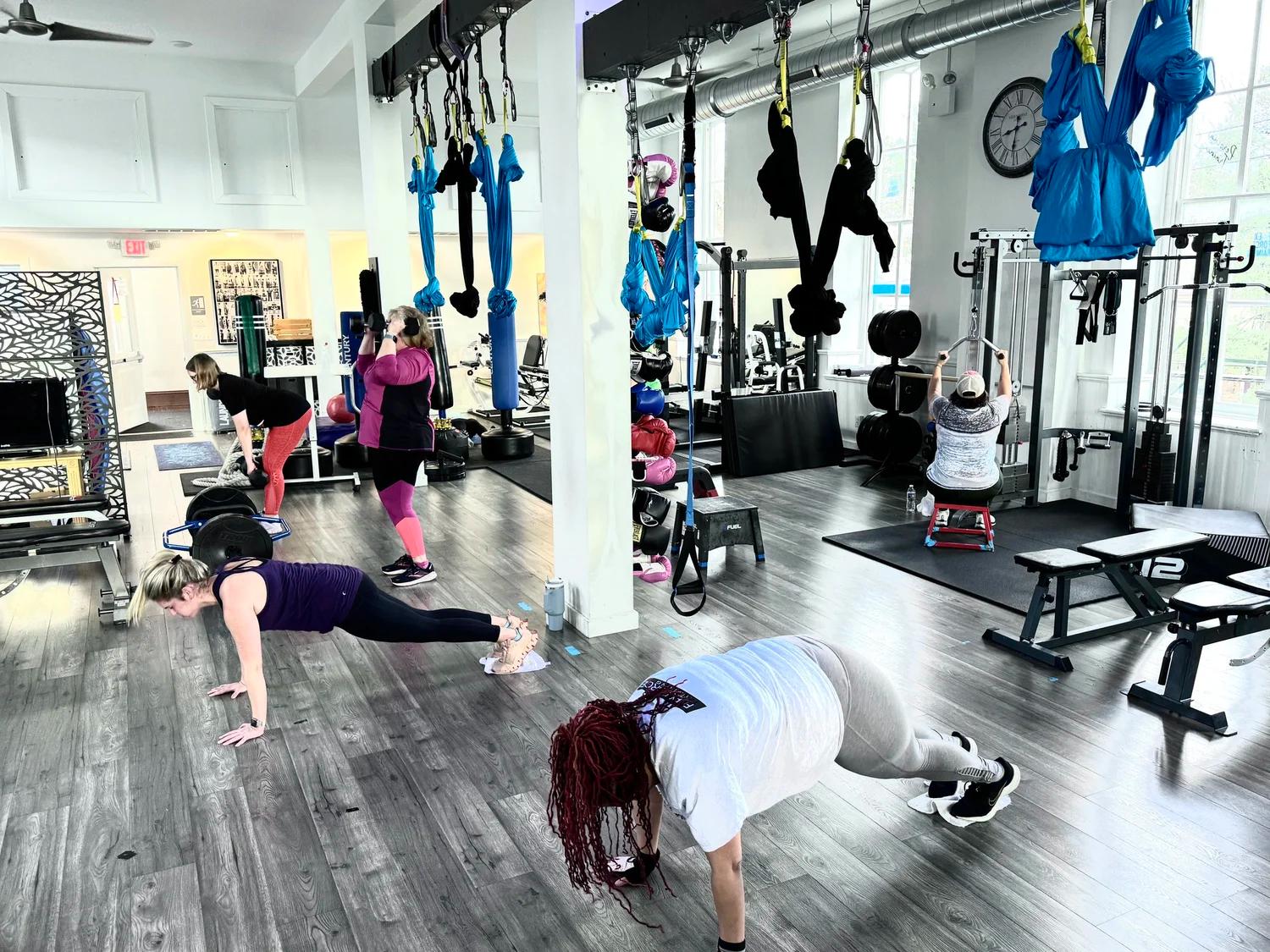Women exercise in a gym, performing planks, deadlifts, and lat pulldowns among other equipment.