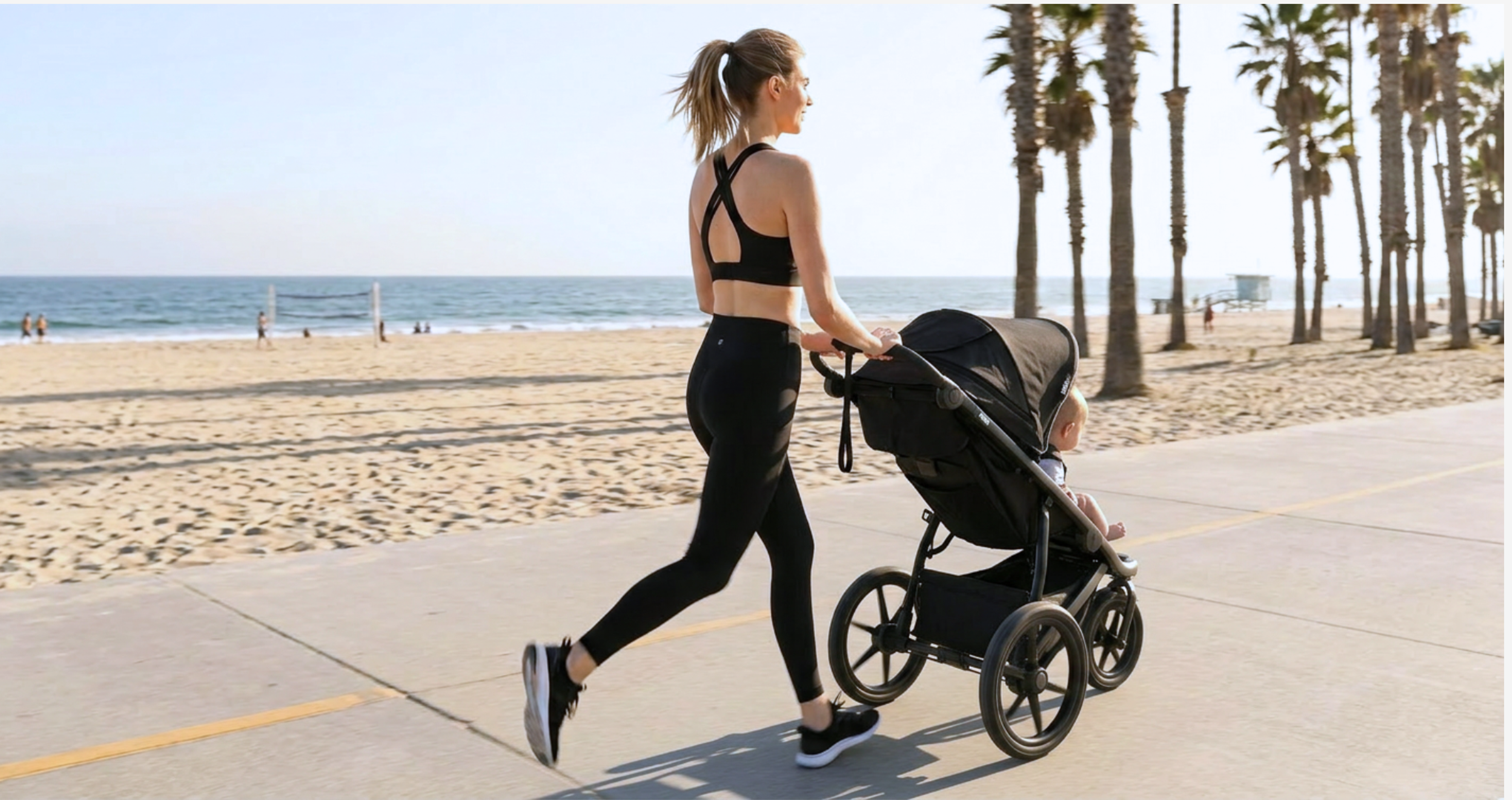 A woman runs along a beach path, pushing a baby in a stroller, with palm trees and the ocean behind her.