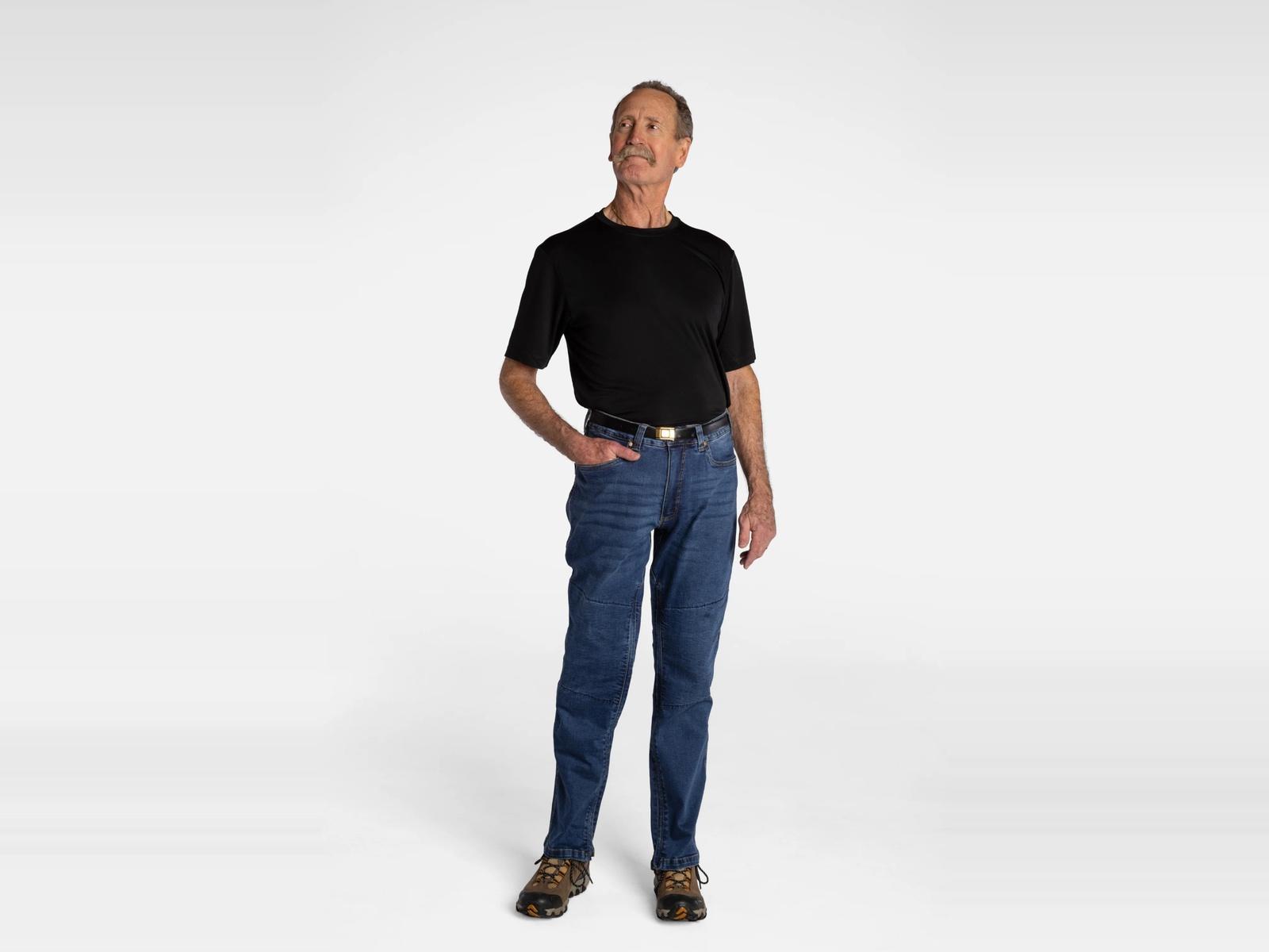 An older man wearing a black t-shirt, blue jeans, and hiking boots stands with one hand in his pocket, looking up and to the left.