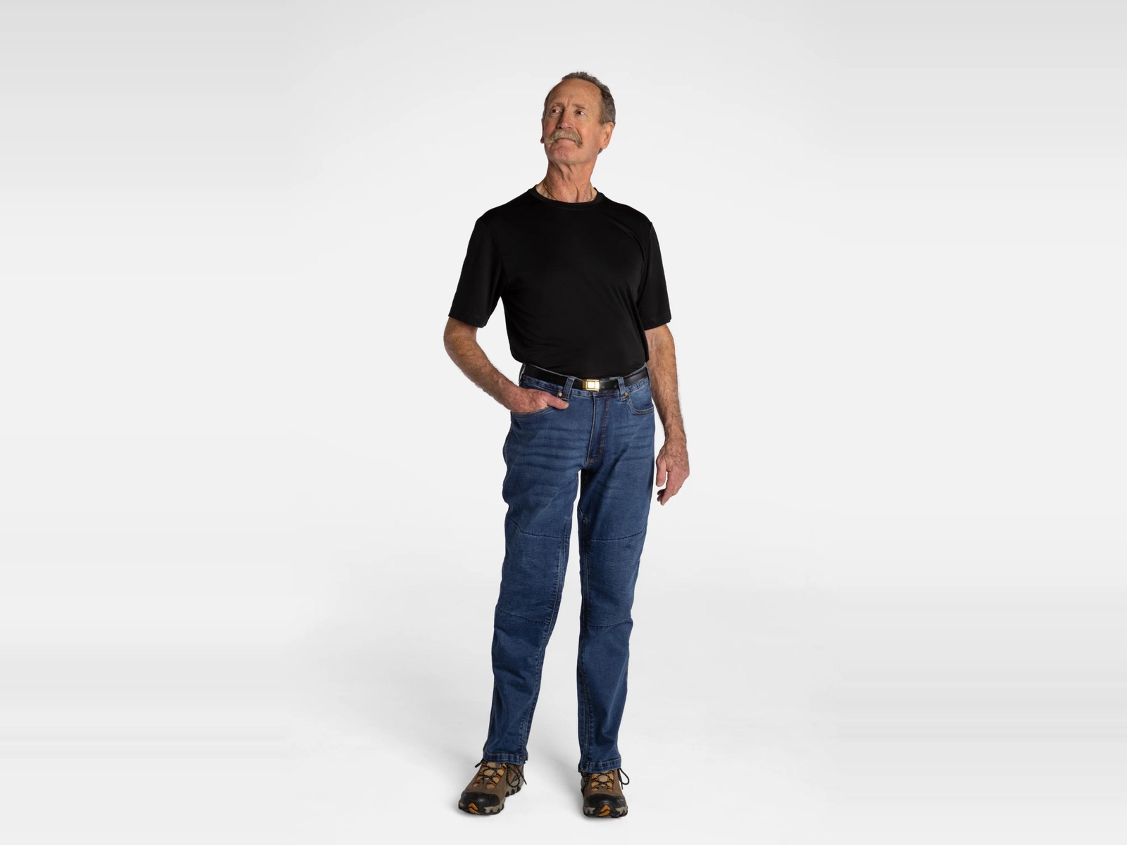 An older man wearing a black t-shirt, blue jeans, and hiking boots stands with one hand in his pocket, looking up and to the left.