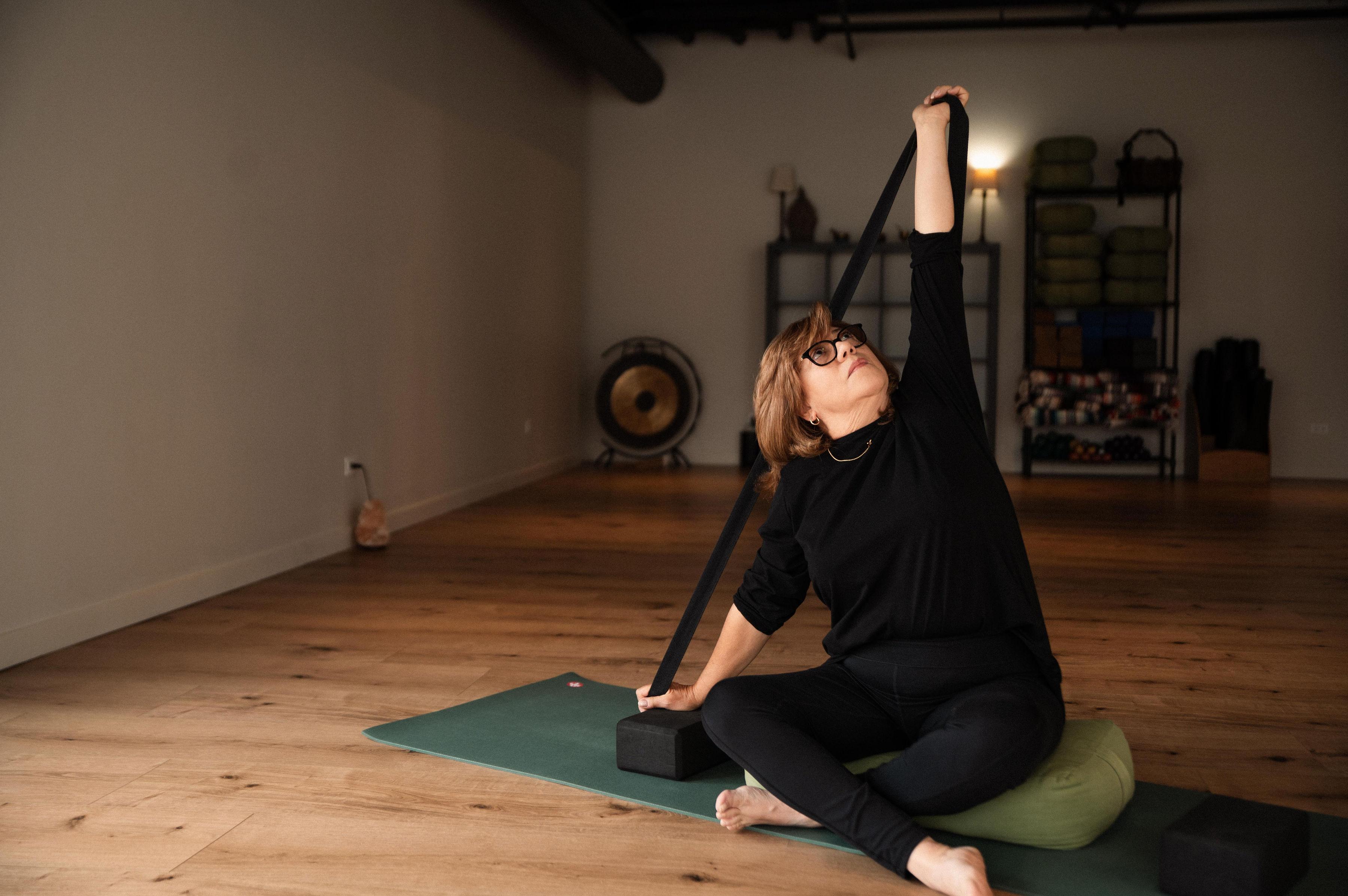 A woman in black does a seated side stretch with a yoga strap and blocks in a studio.