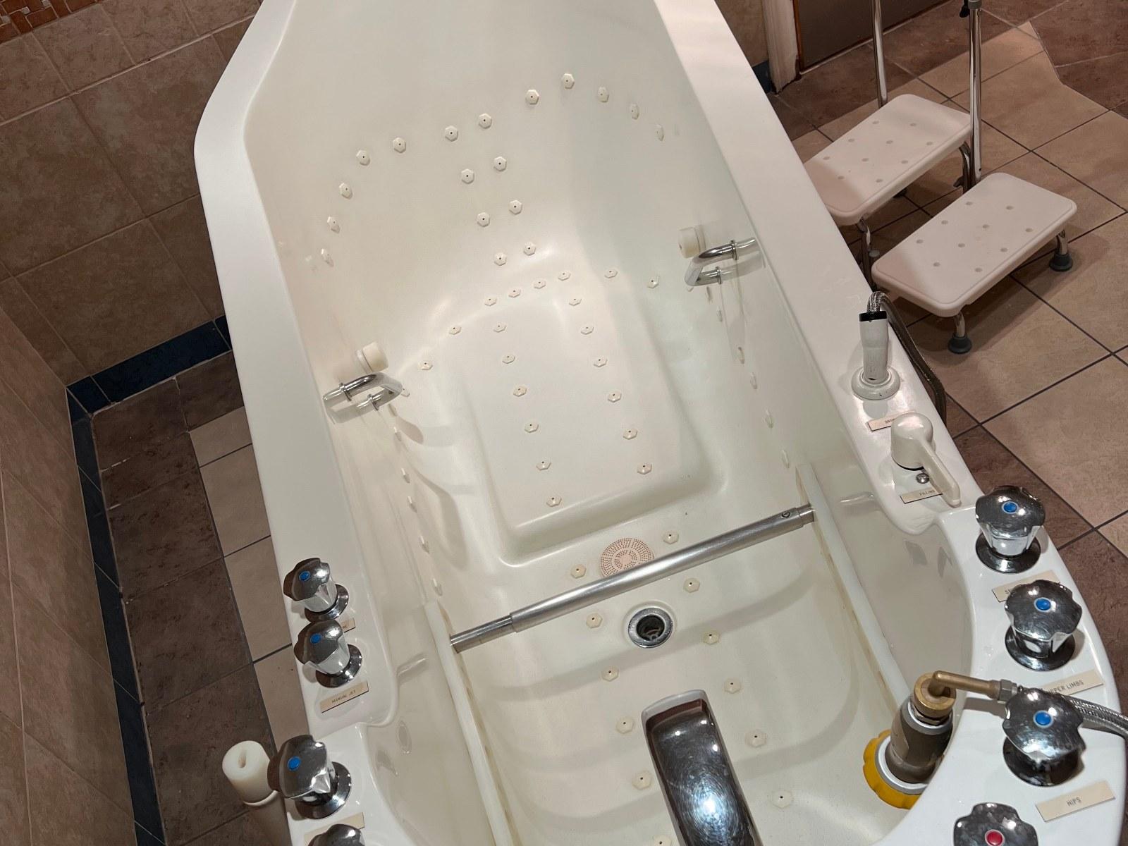 A white therapeutic bathtub with numerous jets, control knobs, an internal grab bar, and two entry steps beside it.