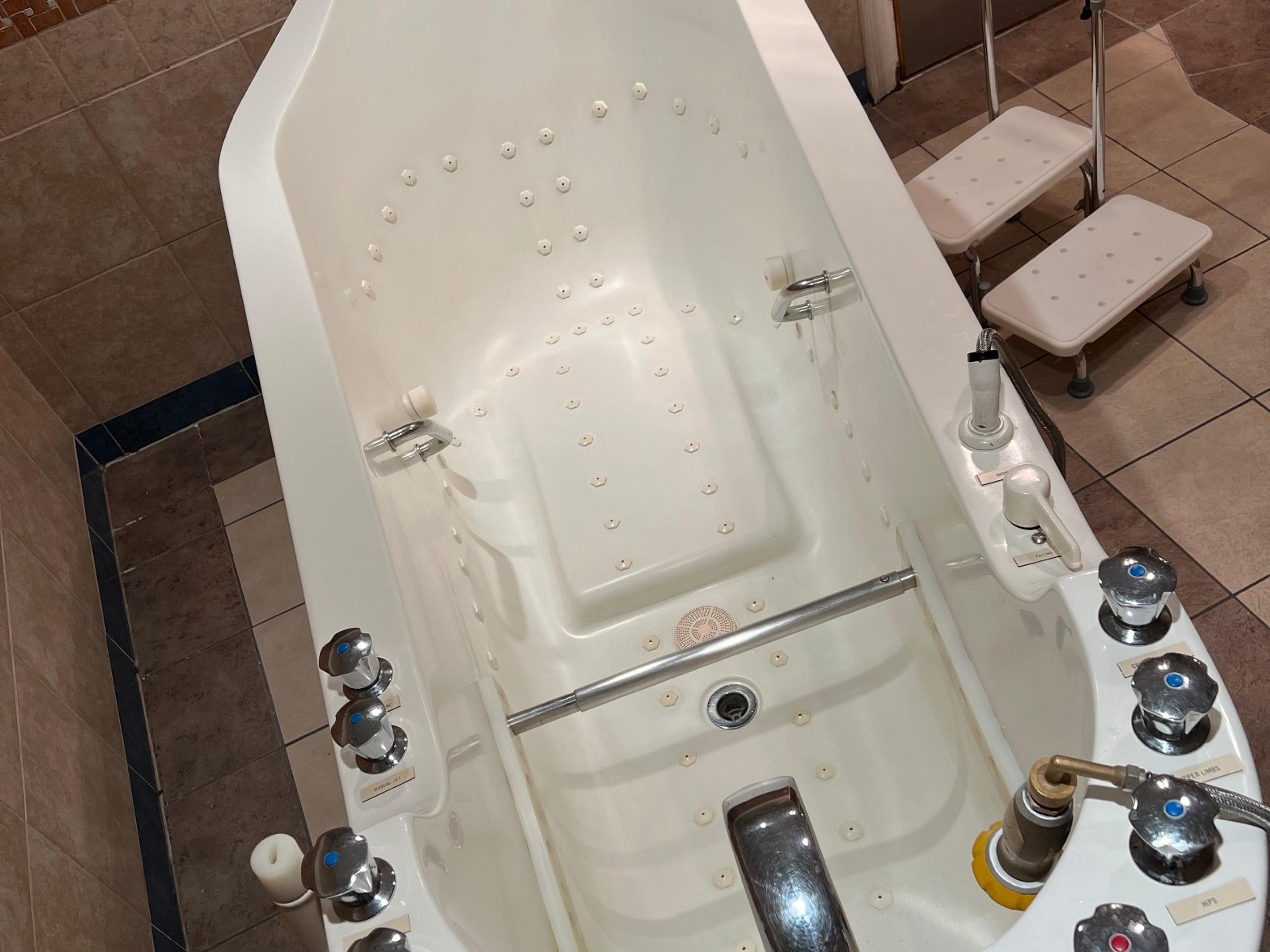 A white therapeutic bathtub with numerous jets, control knobs, an internal grab bar, and two entry steps beside it.
