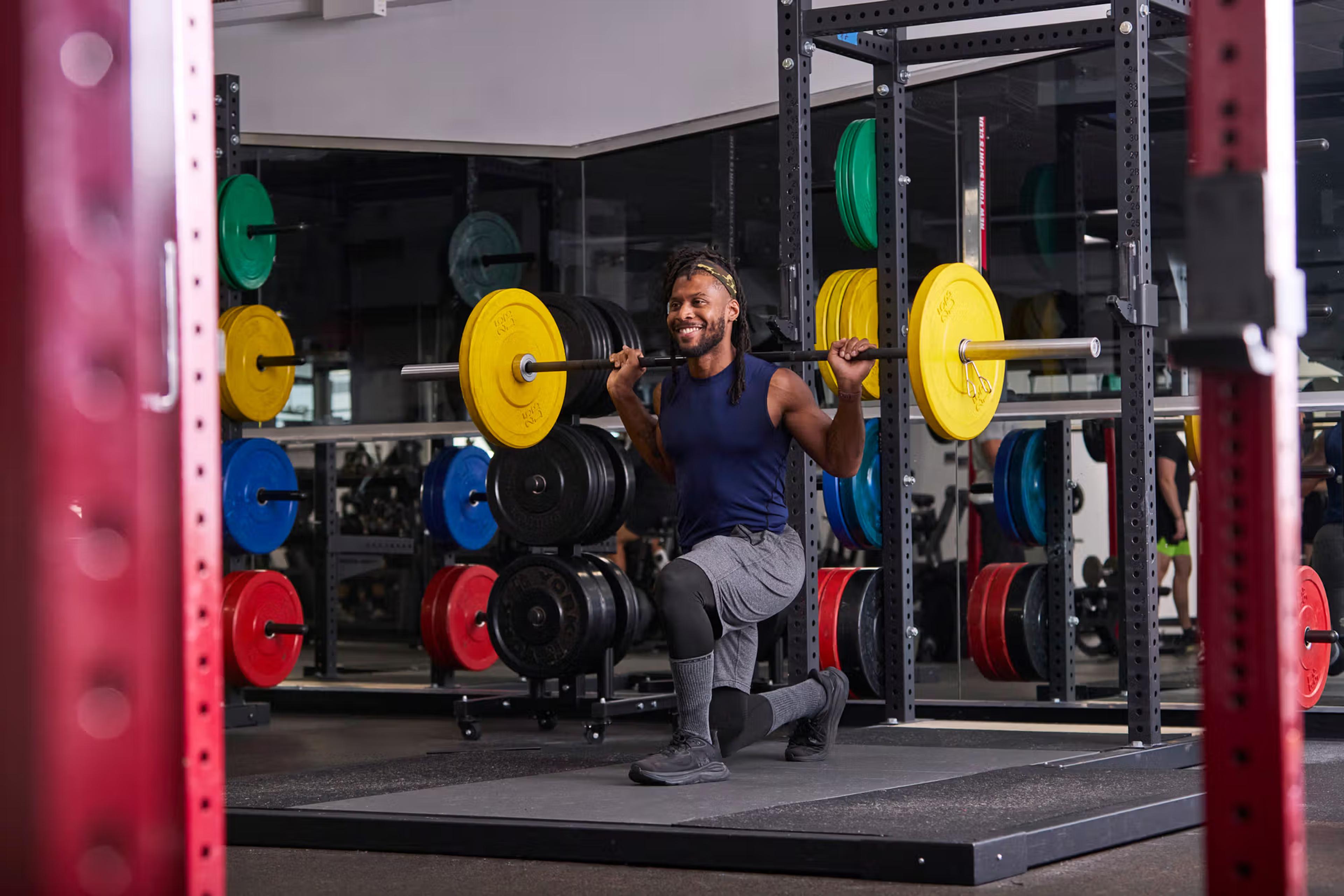 A man smiling while performing a lunge with a barbell on his shoulders in a gym.