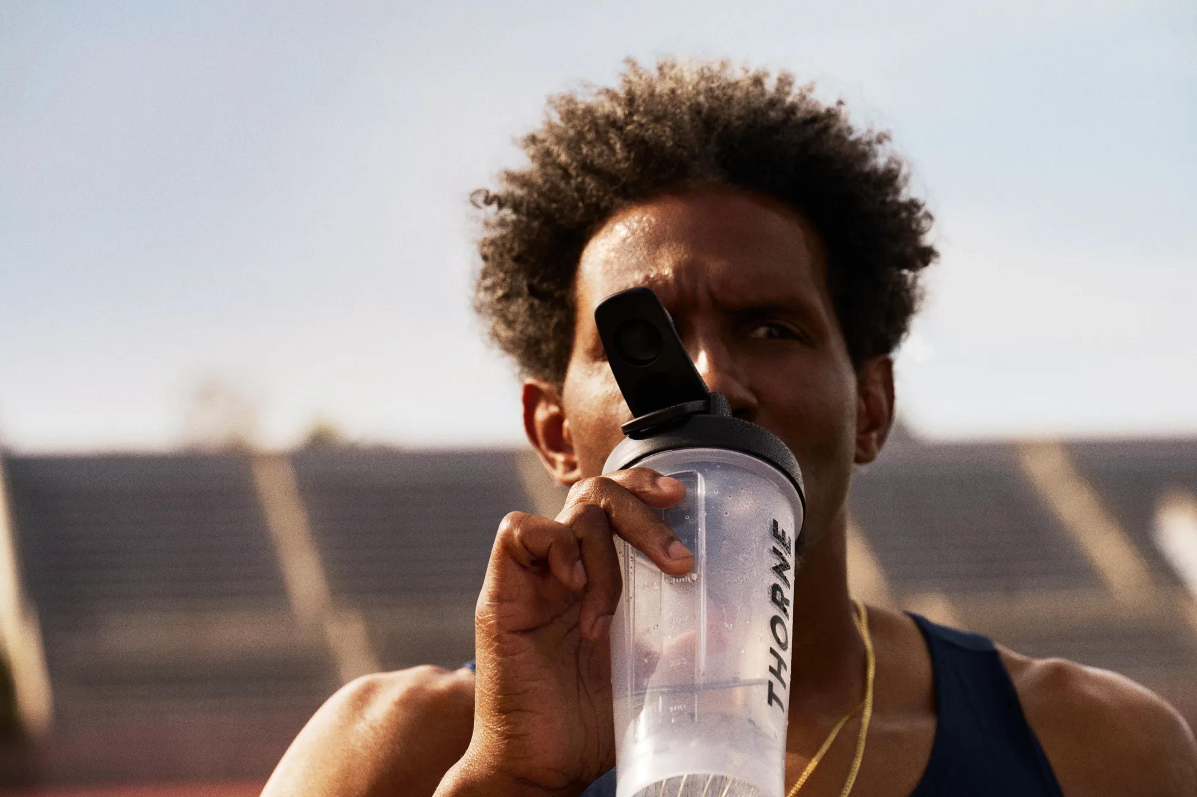 Man with curly hair drinking from a THORNE shaker bottle at a stadium.