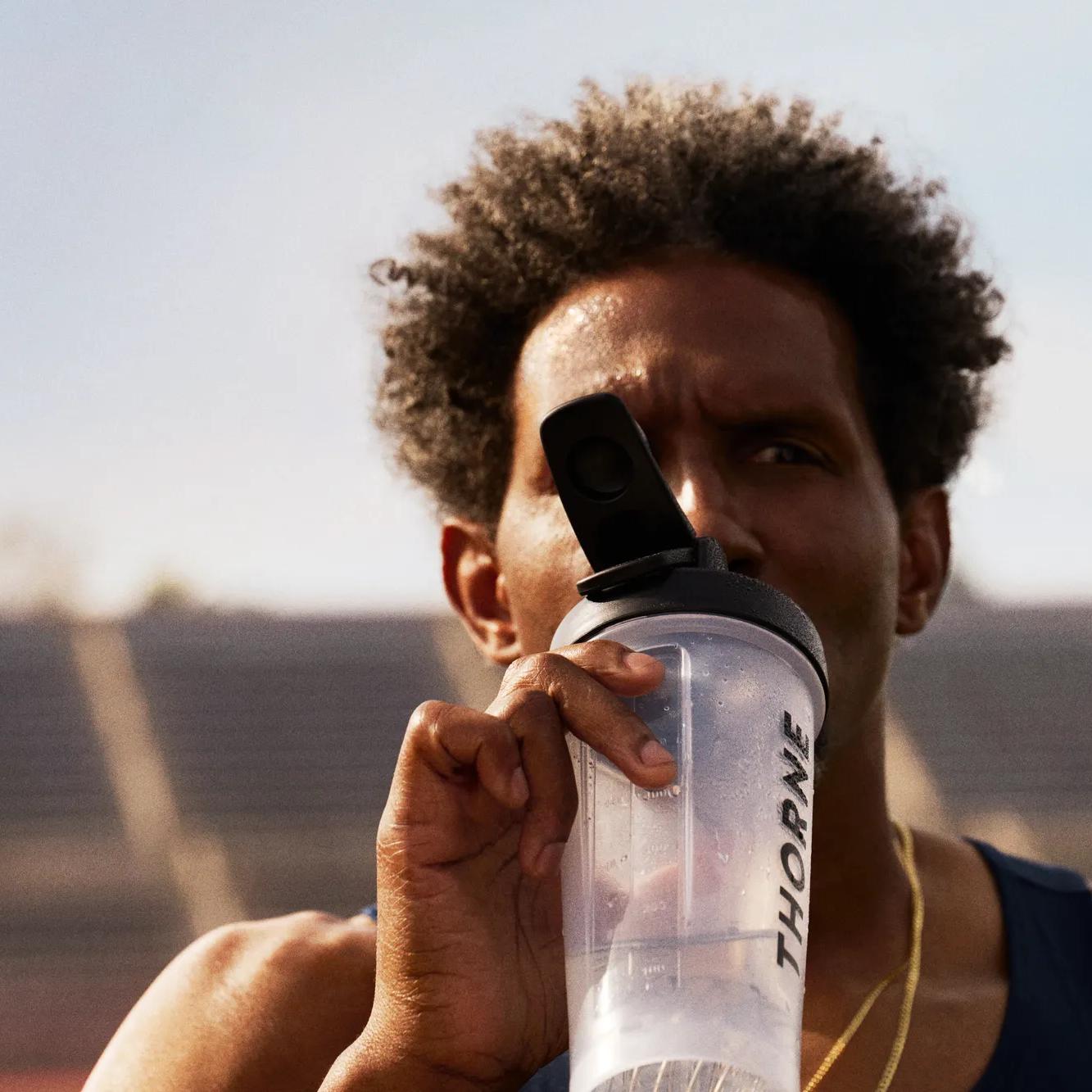 Man with curly hair drinking from a THORNE shaker bottle at a stadium.