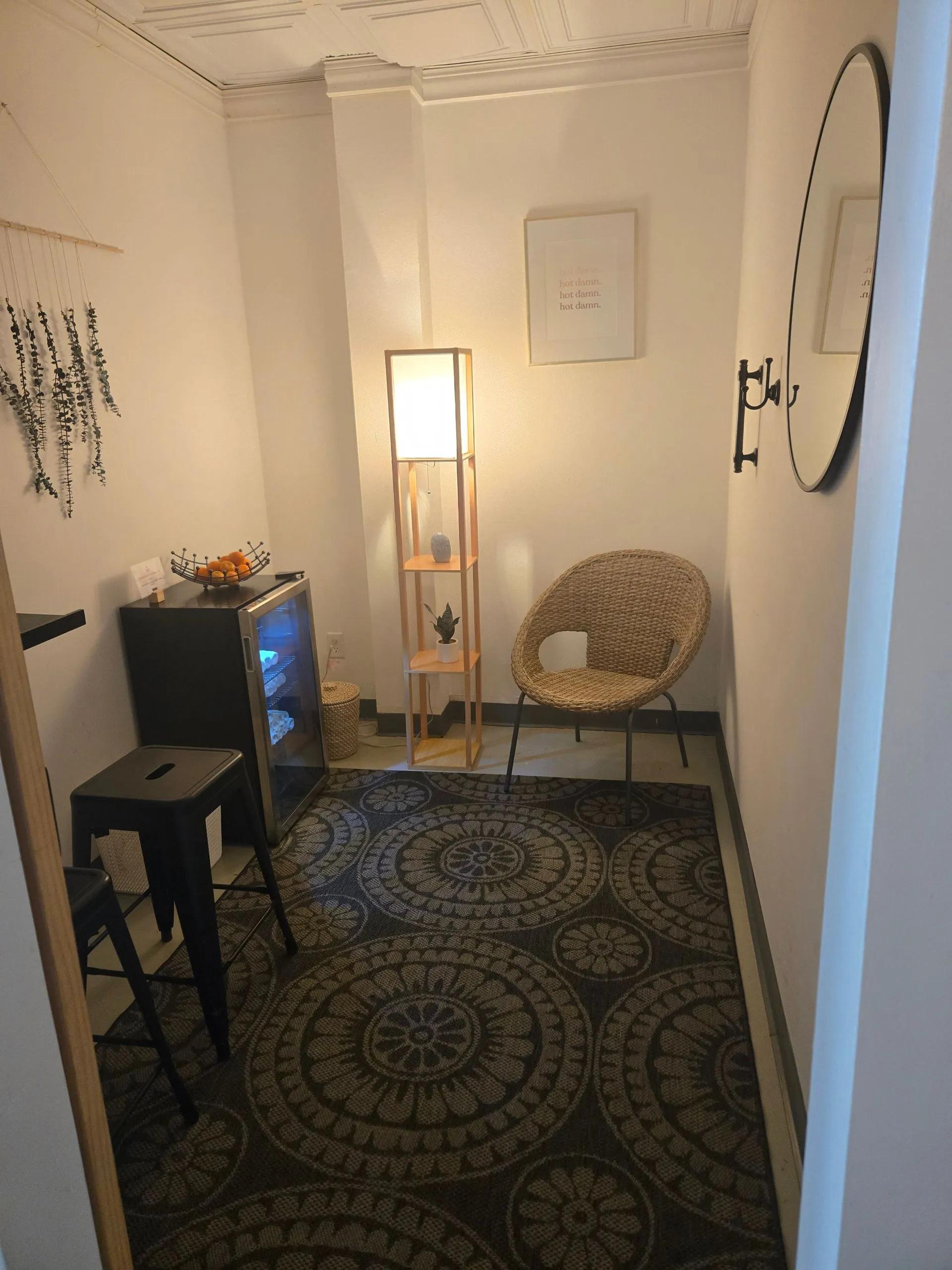 A cozy nook with a patterned rug, mini-fridge, stools, a floor lamp, a rattan chair, and a large oval mirror on the wall.