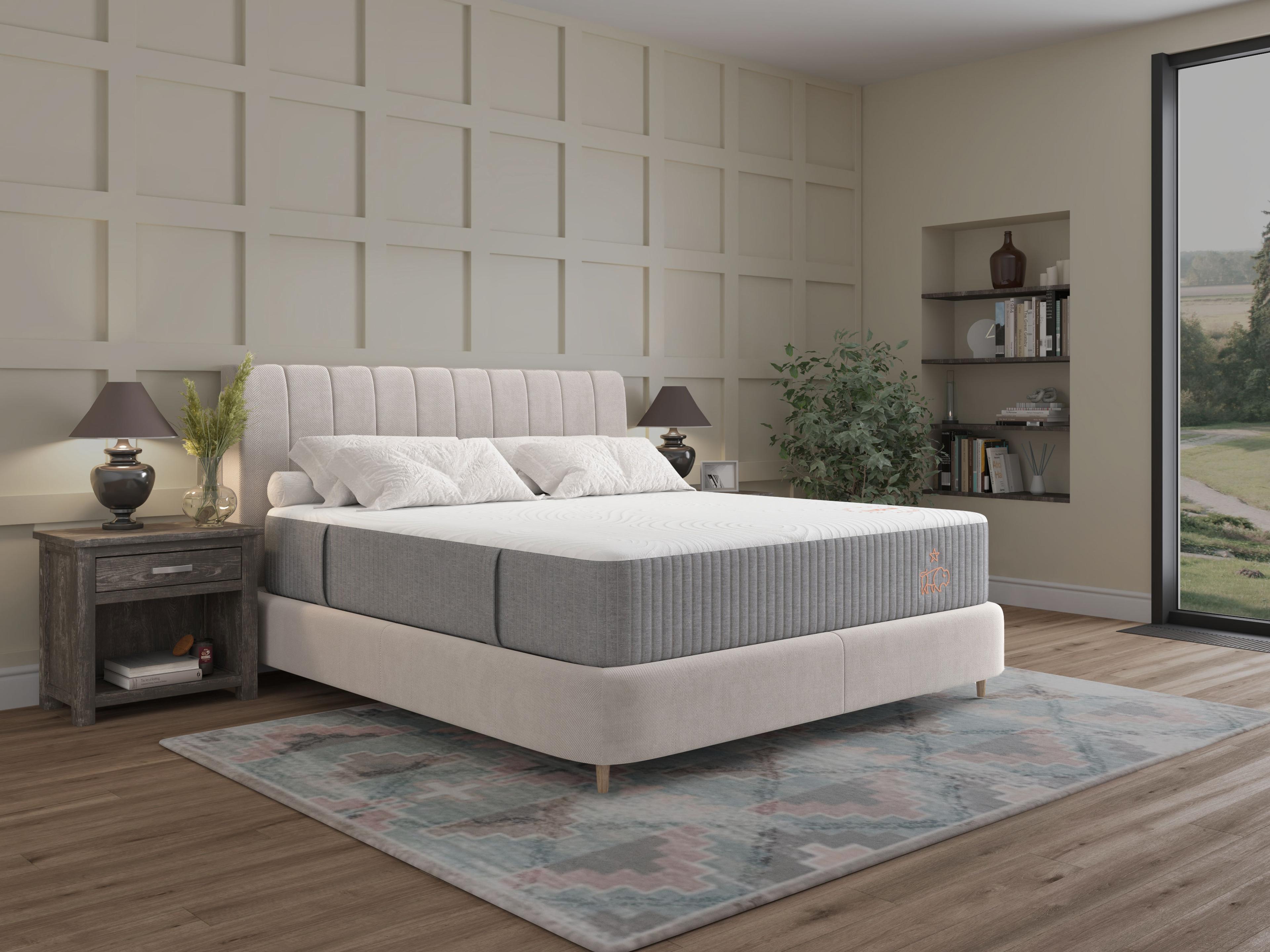 A bedroom features a bed with a white and grey mattress, an upholstered headboard, a nightstand with a lamp, and a patterned rug, with a large window overlooking a garden.