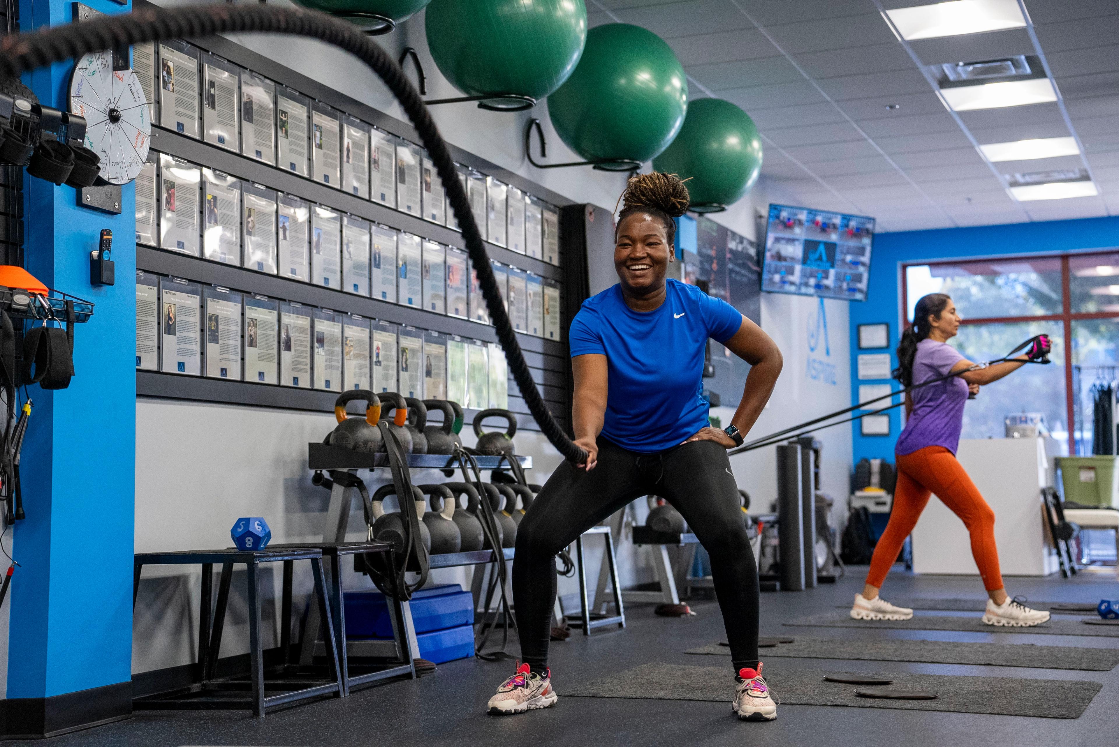 A smiling Black woman trains with battle ropes in a gym, while another woman exercises with a resistance band in the background.