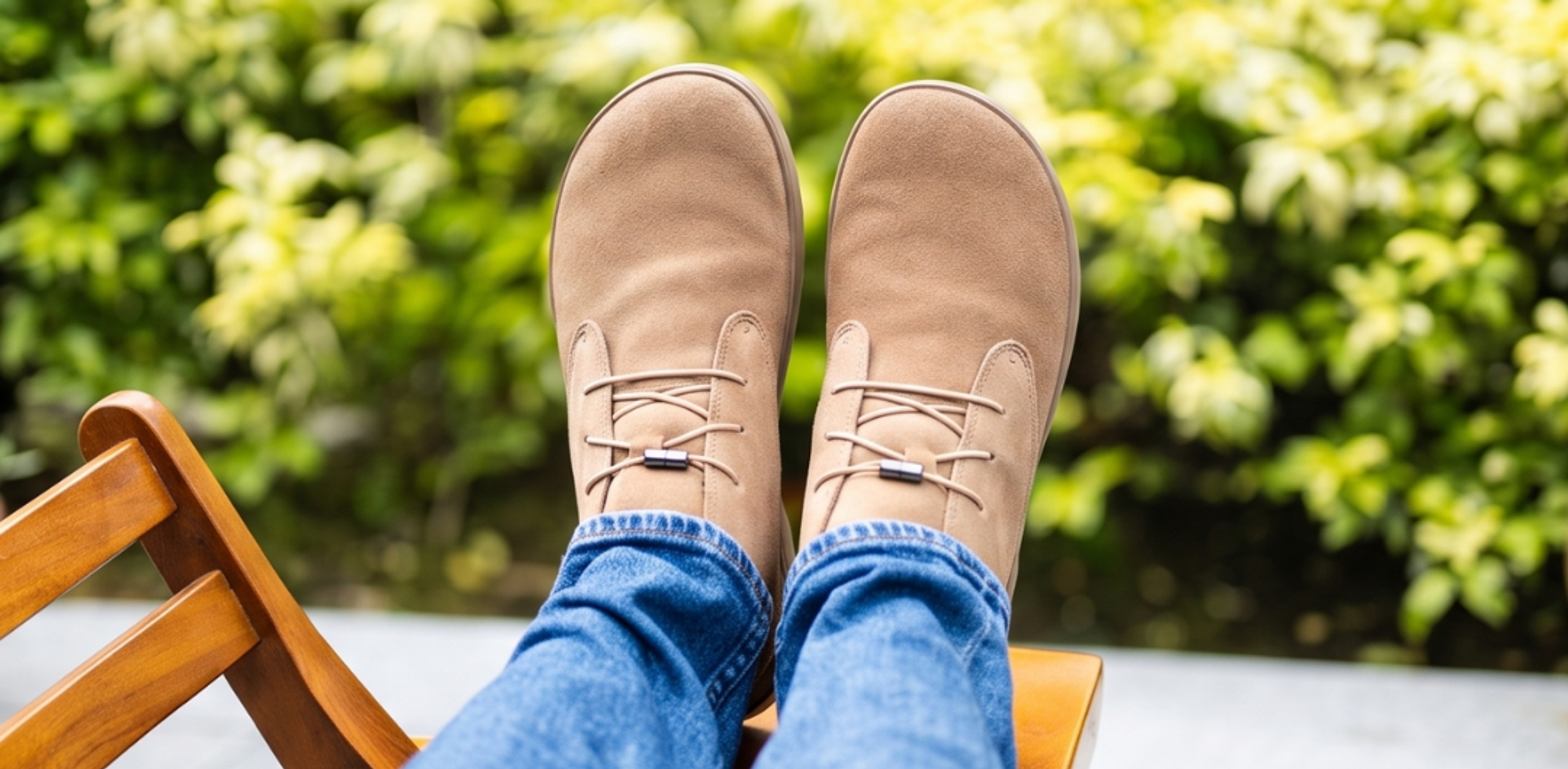 Overhead view of feet in tan minimalist shoes and blue jeans, resting on a wooden chair with a blurry green background.
