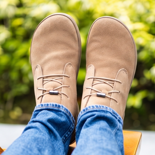Overhead view of feet in tan minimalist shoes and blue jeans, resting on a wooden chair with a blurry green background.