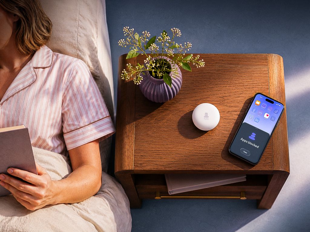 A person reads in bed next to a nightstand with a white circular device and a smartphone displaying "Apps blocked."