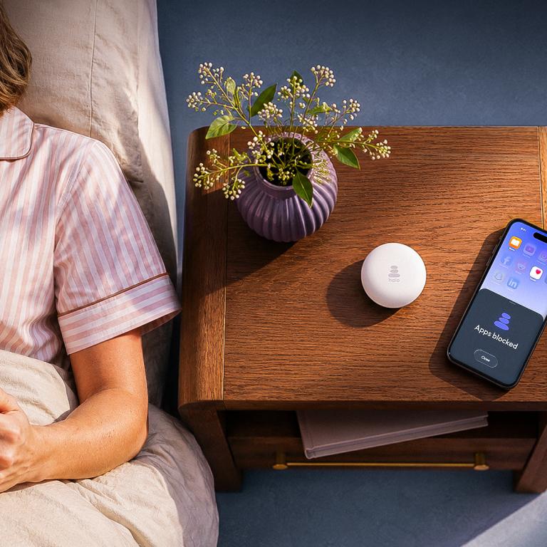 A person reads in bed next to a nightstand with a white circular device and a smartphone displaying "Apps blocked."