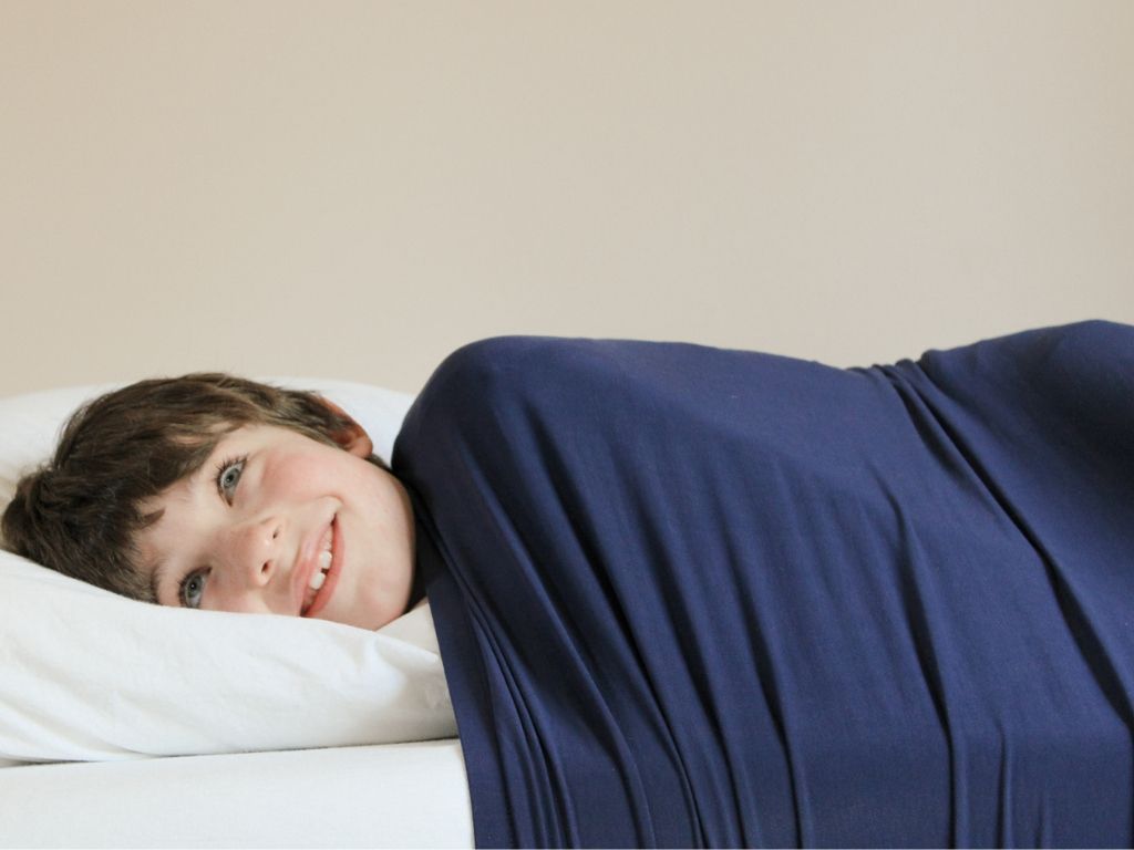 Smiling boy with his head on a pillow, body covered by Cocooner's dark blue compression sheet.
