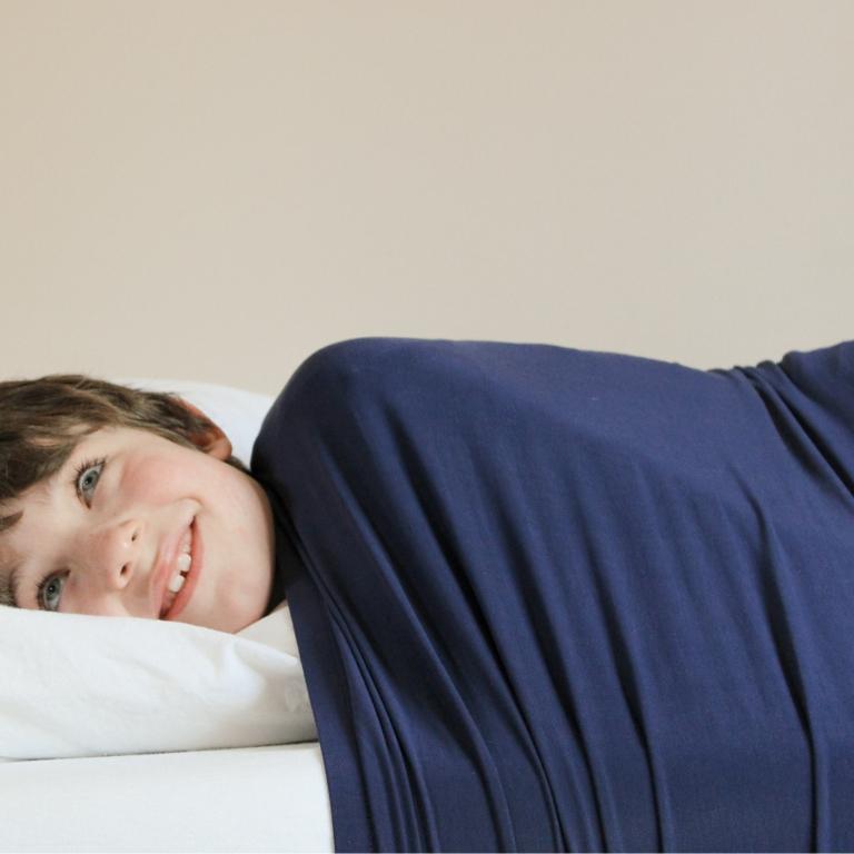 Smiling boy with his head on a pillow, body covered by Cocooner's dark blue compression sheet.