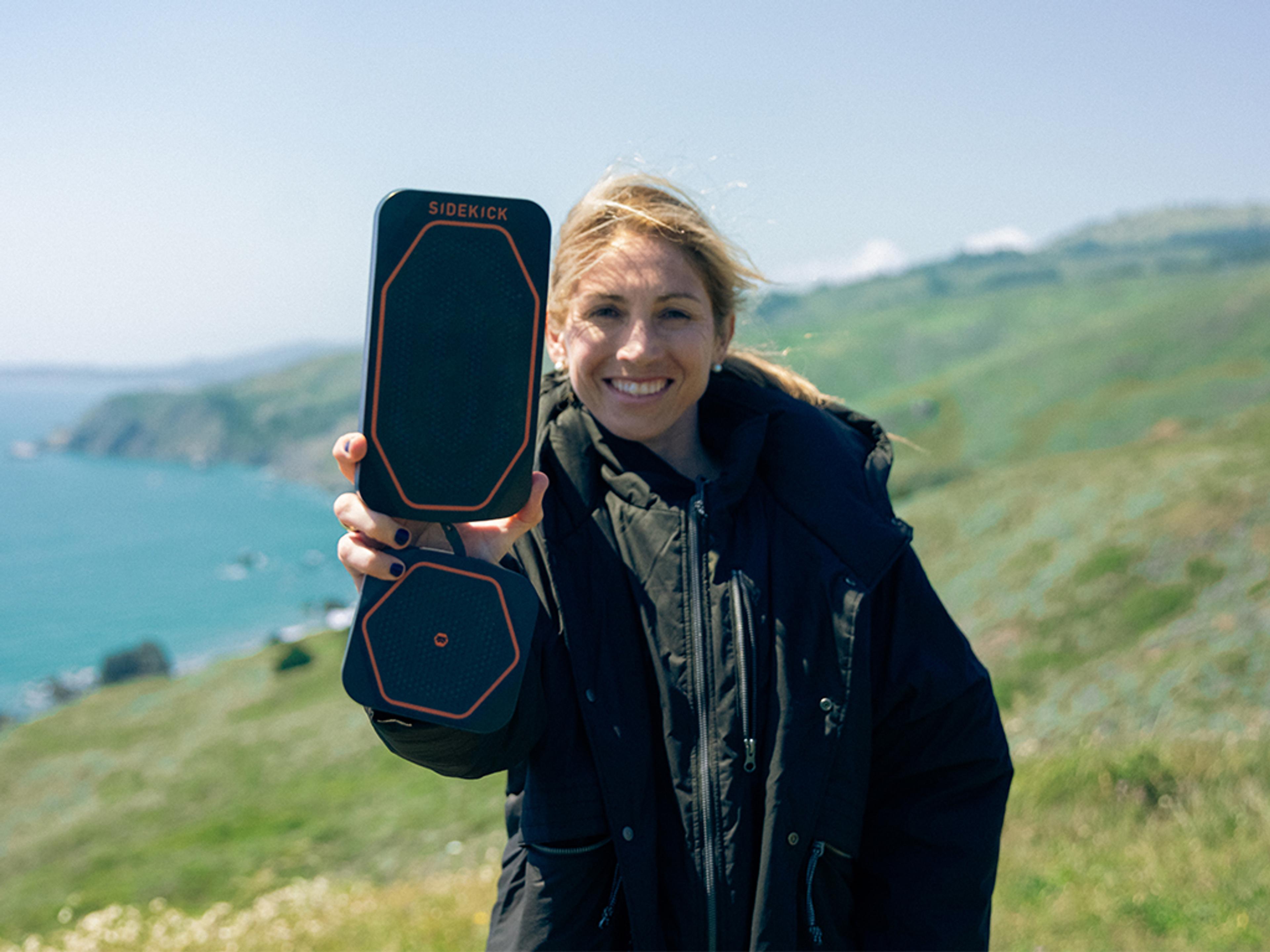 A smiling woman holds up two black "Sidekick" devices, one rectangular and one hexagonal, on a sunny hillside overlooking the ocean.