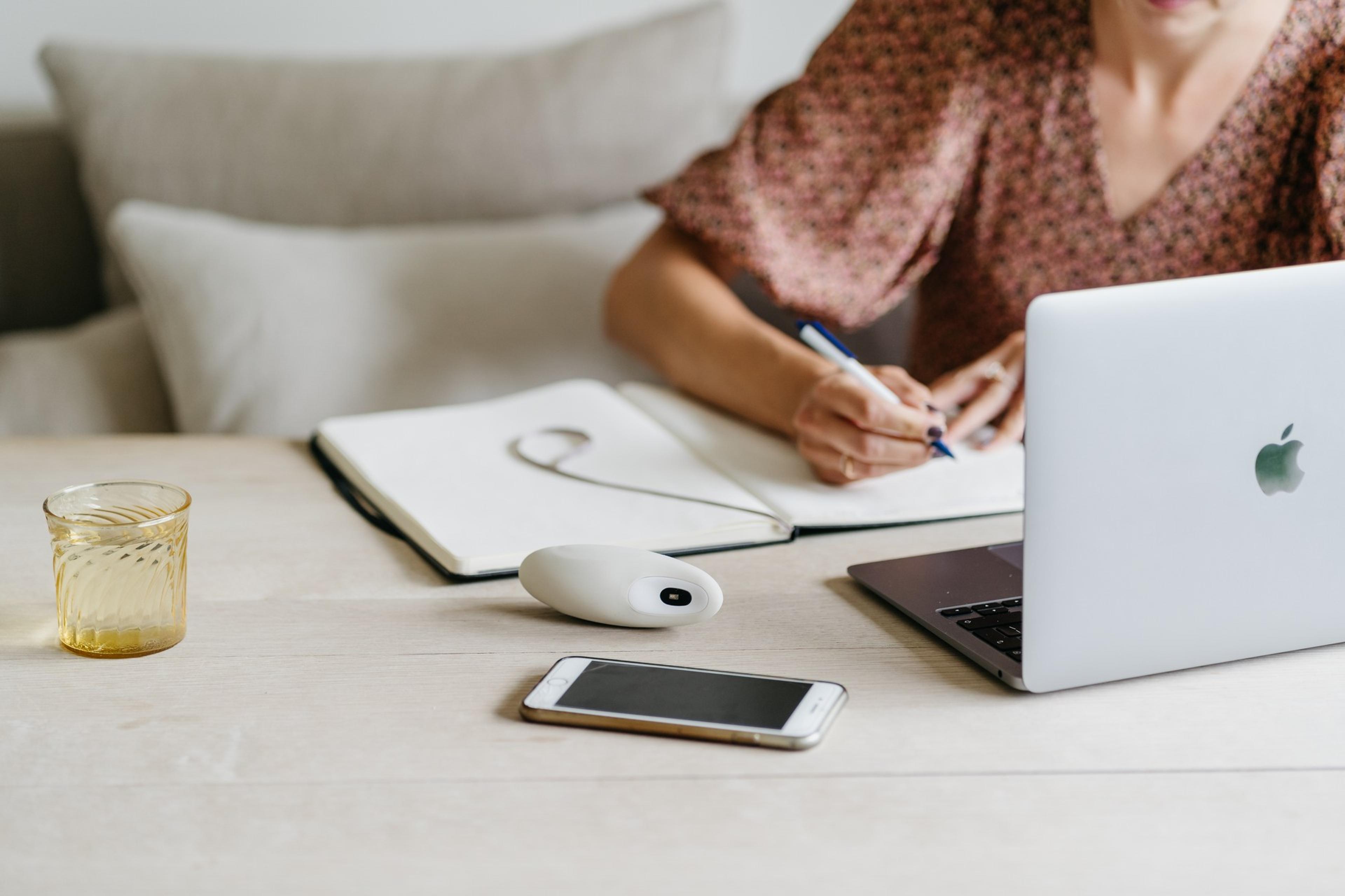 A person writes in a notebook at a desk with a moonbird, laptop, mouse, and phone.