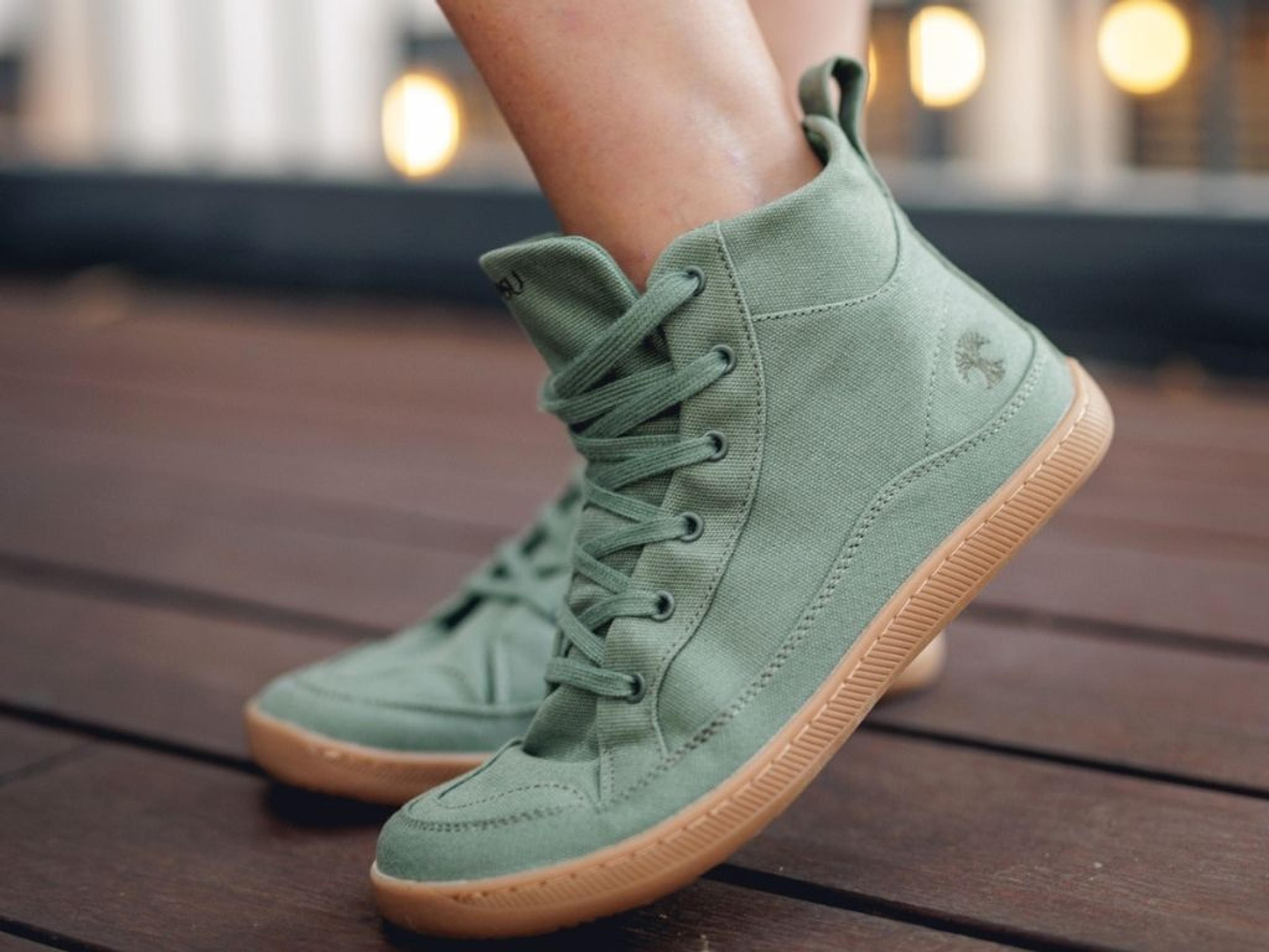 Olive green canvas high-top sneakers with a tree logo and gum soles, worn by a person on a wooden deck.