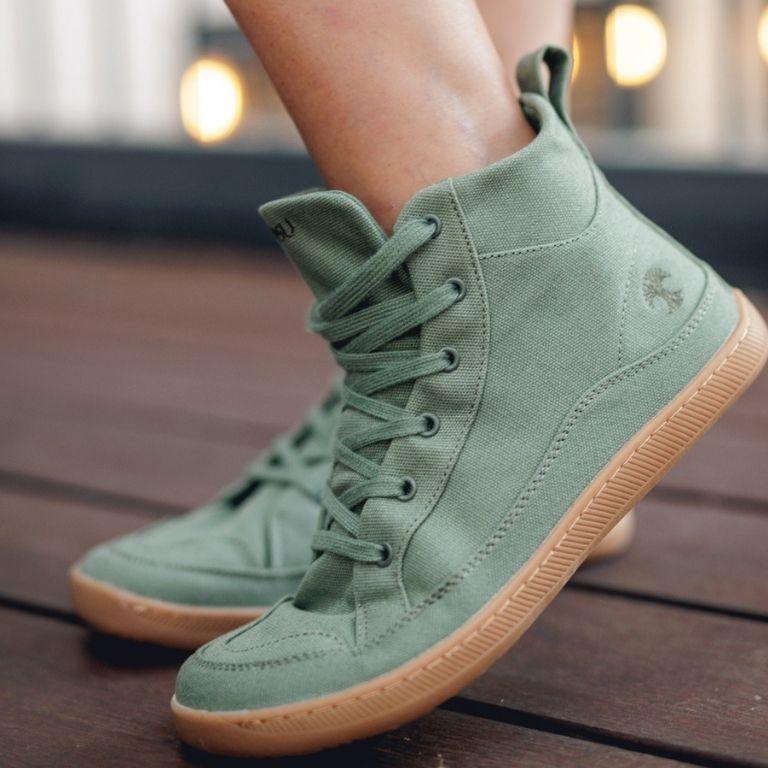 Olive green canvas high-top sneakers with a tree logo and gum soles, worn by a person on a wooden deck.
