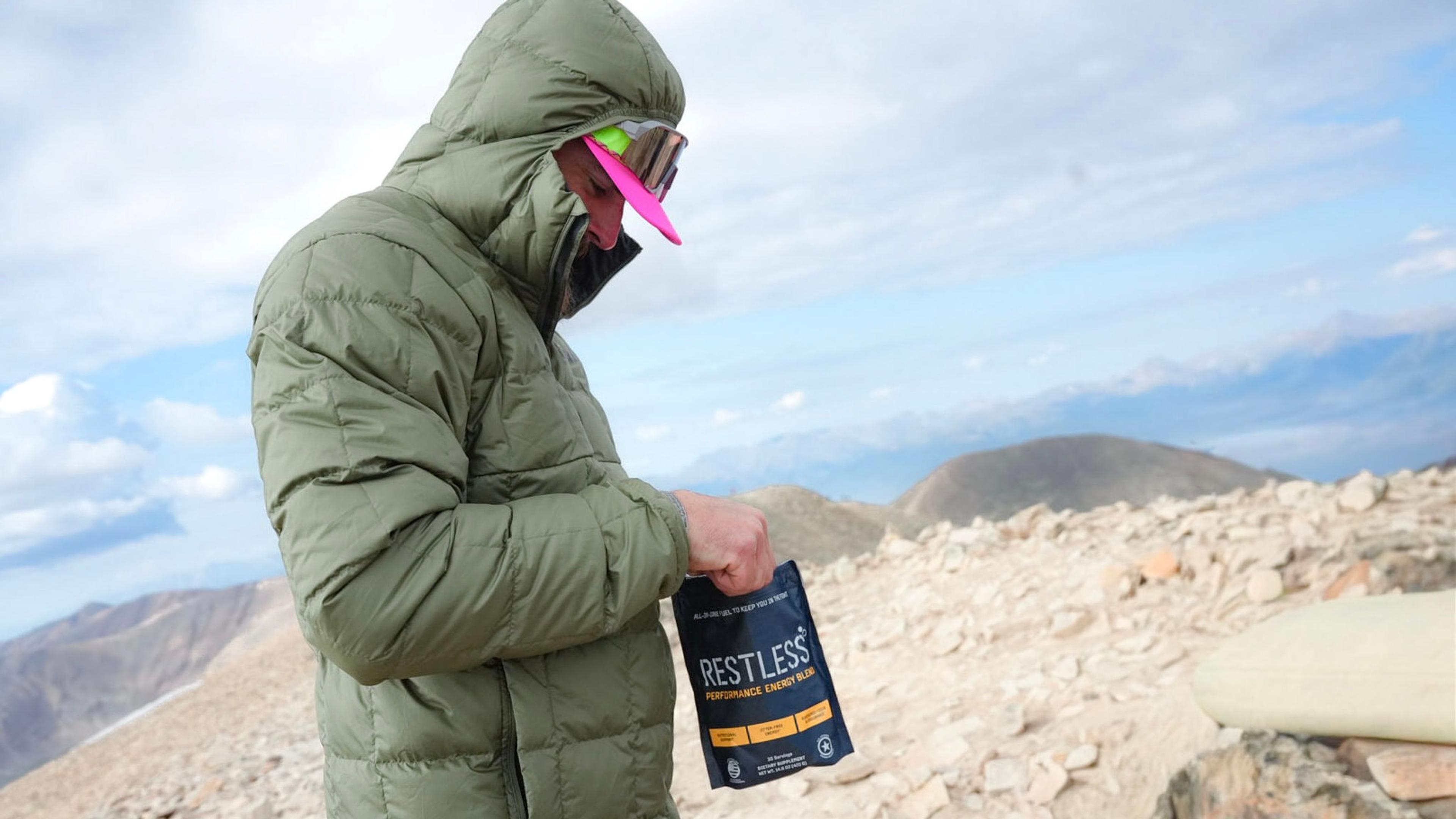 Hiker in a green puffer jacket and pink-brimmed goggles holds a "Restless Performance Energy Mix" pouch on a rocky mountain peak.