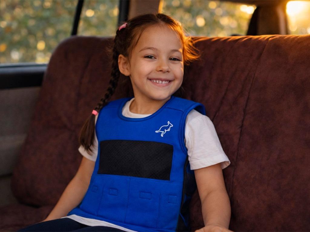 A smiling young girl in a blue vest with a kangaroo logo sits in a car.