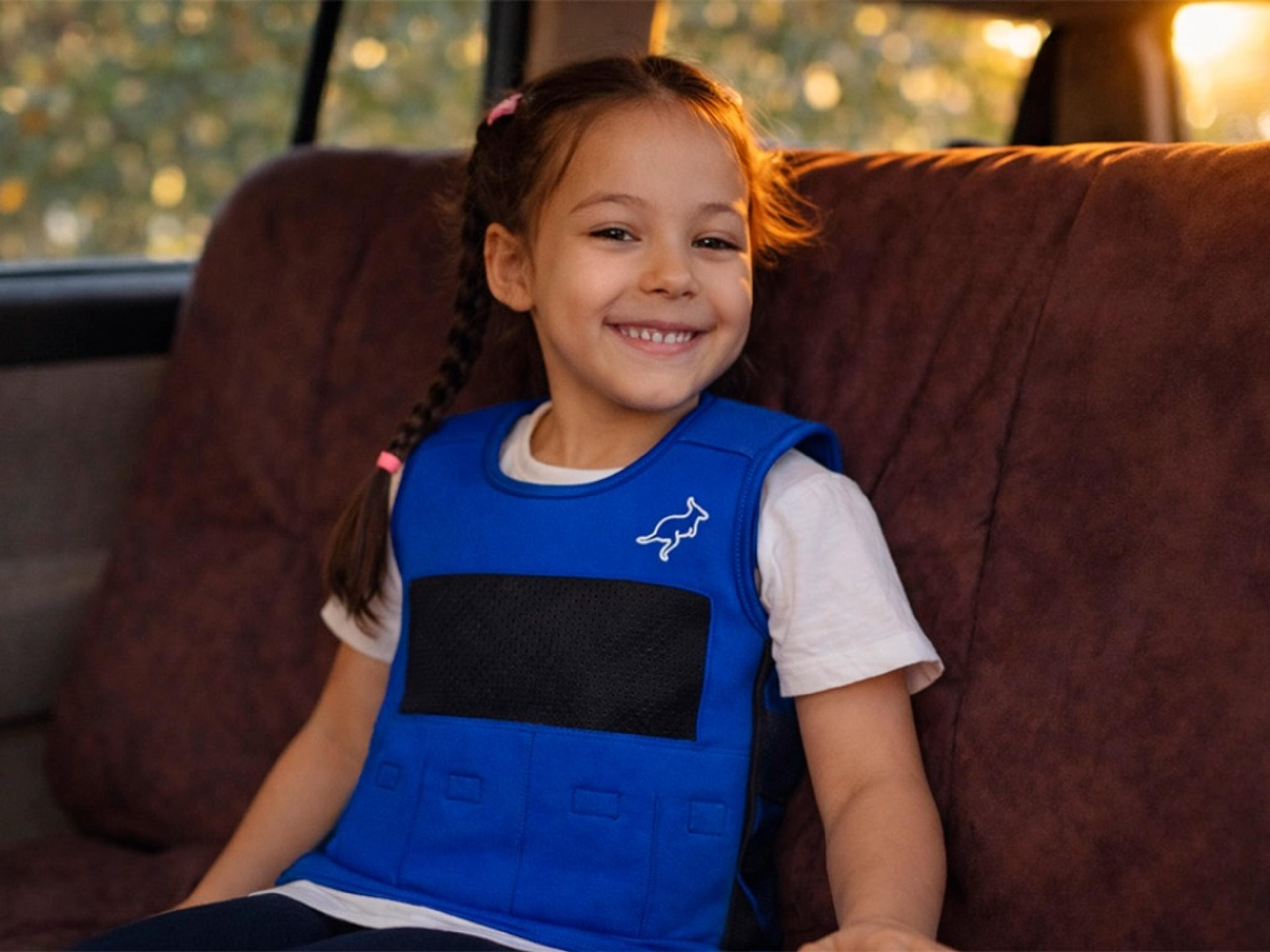 A smiling young girl in a blue vest with a kangaroo logo sits in a car.
