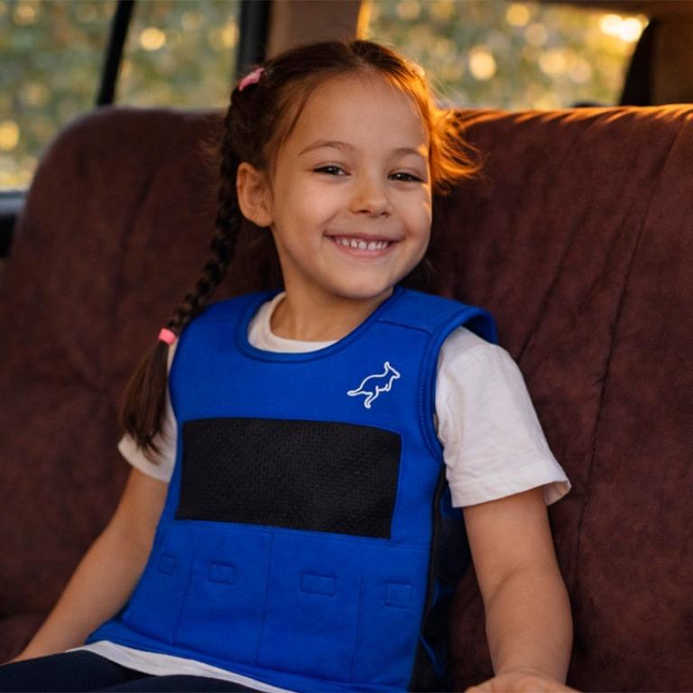 A smiling young girl in a blue vest with a kangaroo logo sits in a car.