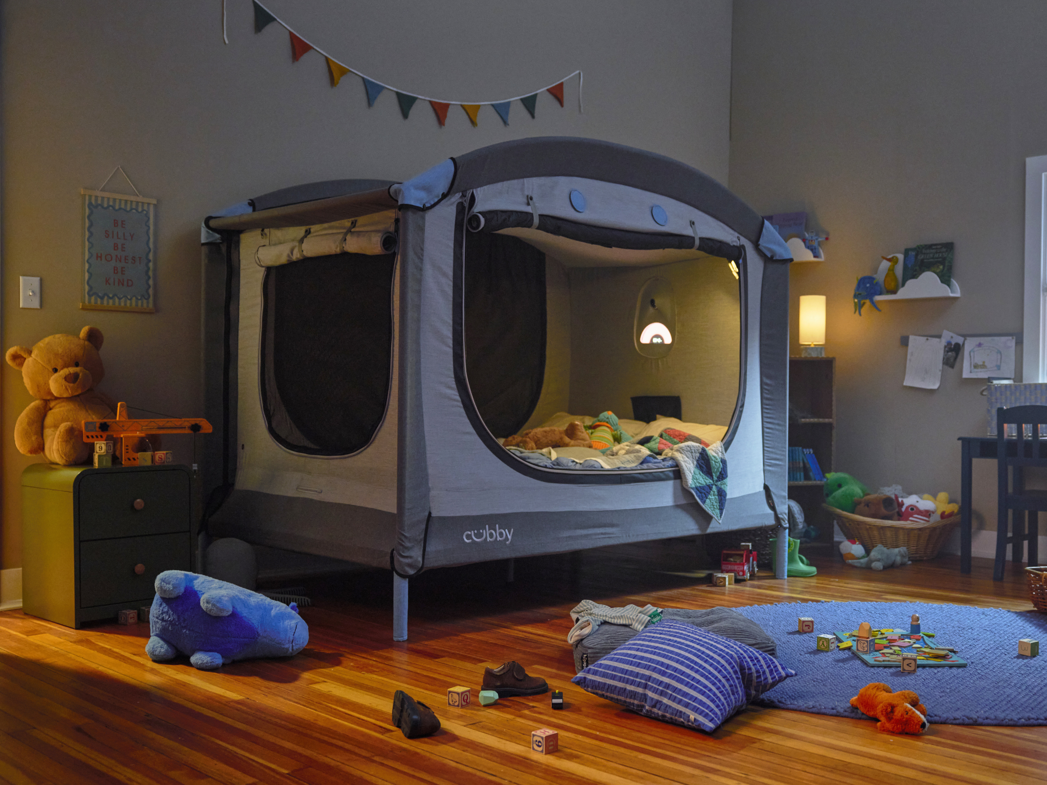 A child's bedroom featuring a grey and blue cubby bed enclosure, lit from within, surrounded by toys on a wooden floor.