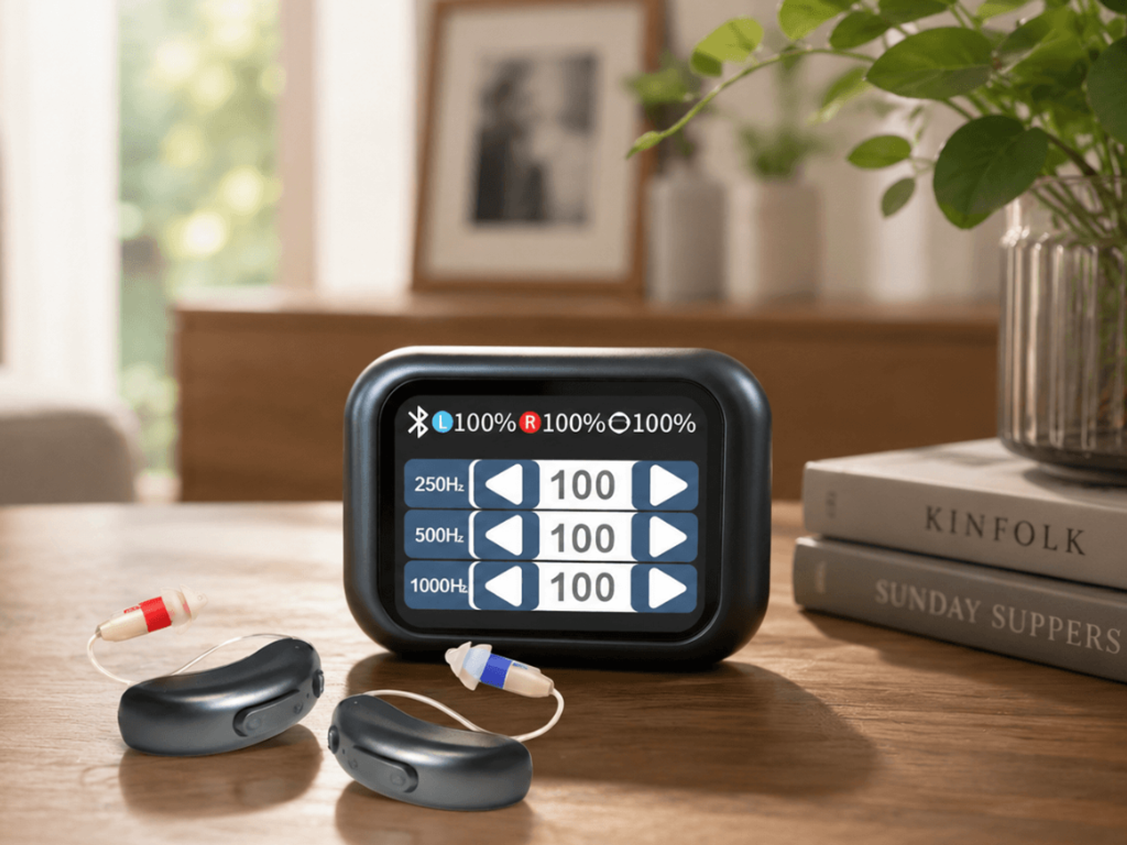 A black digital hearing aid controller displays audio settings, next to two dark gray hearing aids with colored tips on a wooden table.